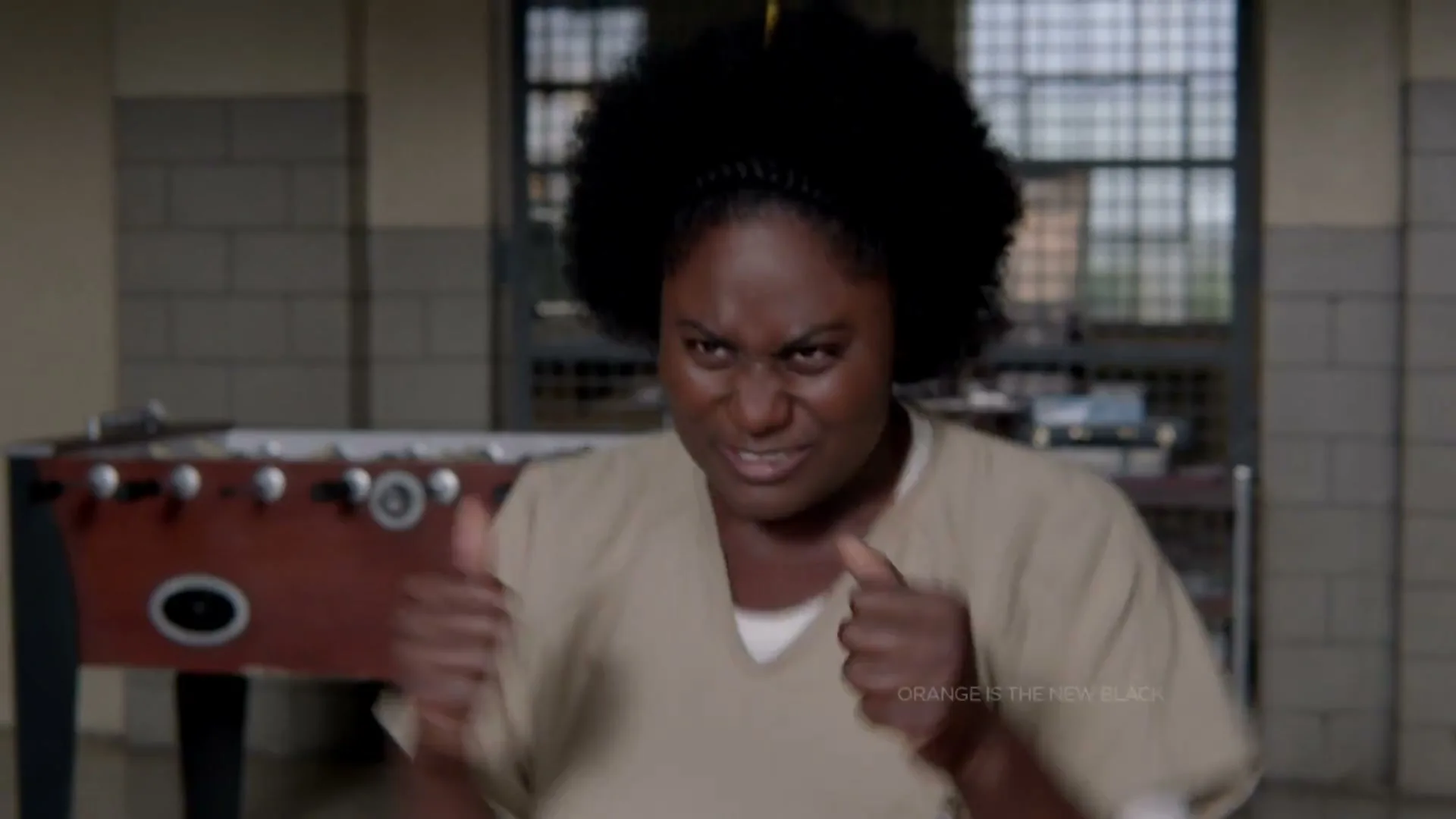 A Black woman with an afro and a pencil in her hair, wearing a prison uniform, makes a funny face and gestures with her hands in a prison common room.