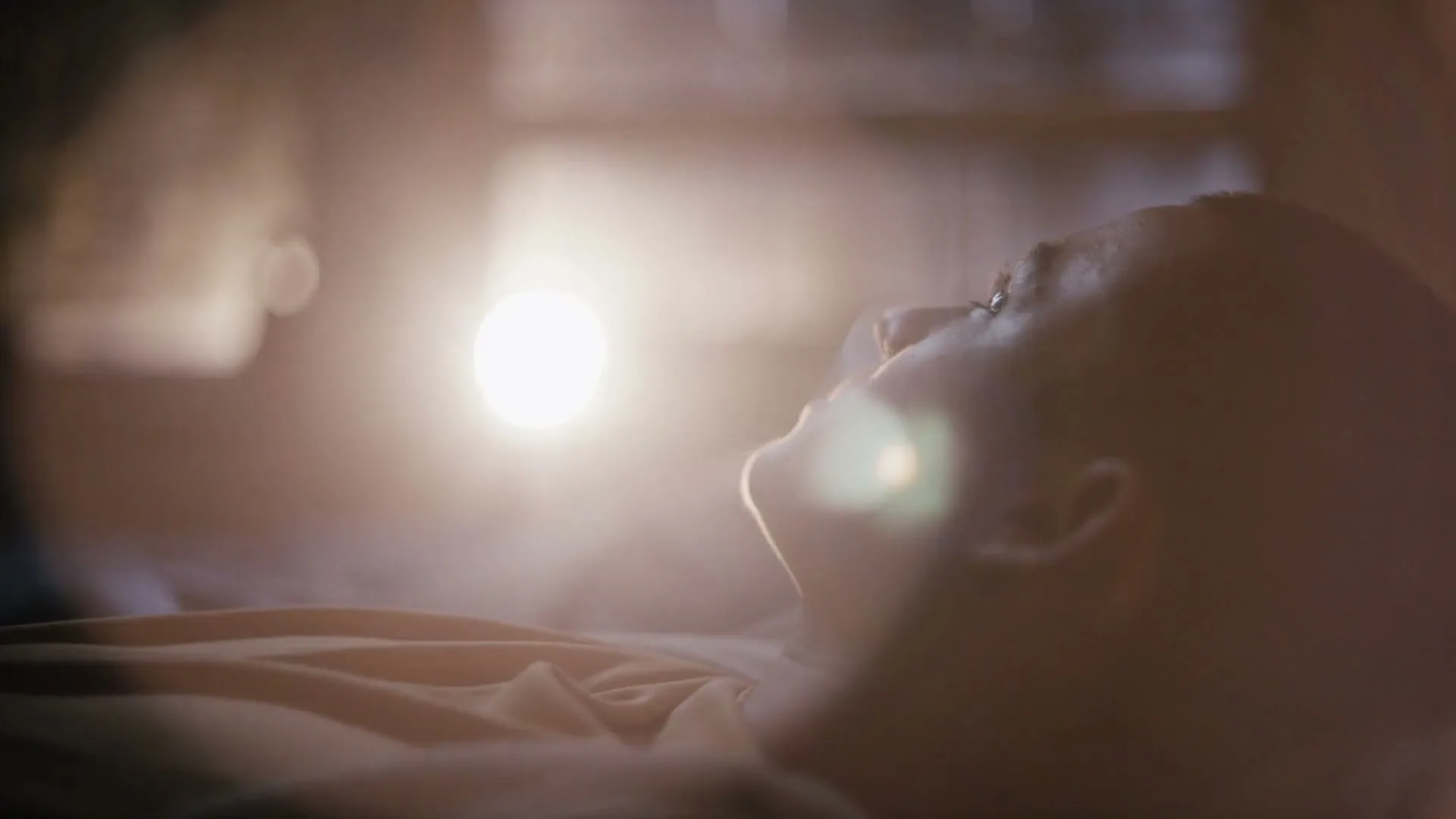 A young person lies in bed, looking up at a bright light source, possibly a phone, in a dark room.