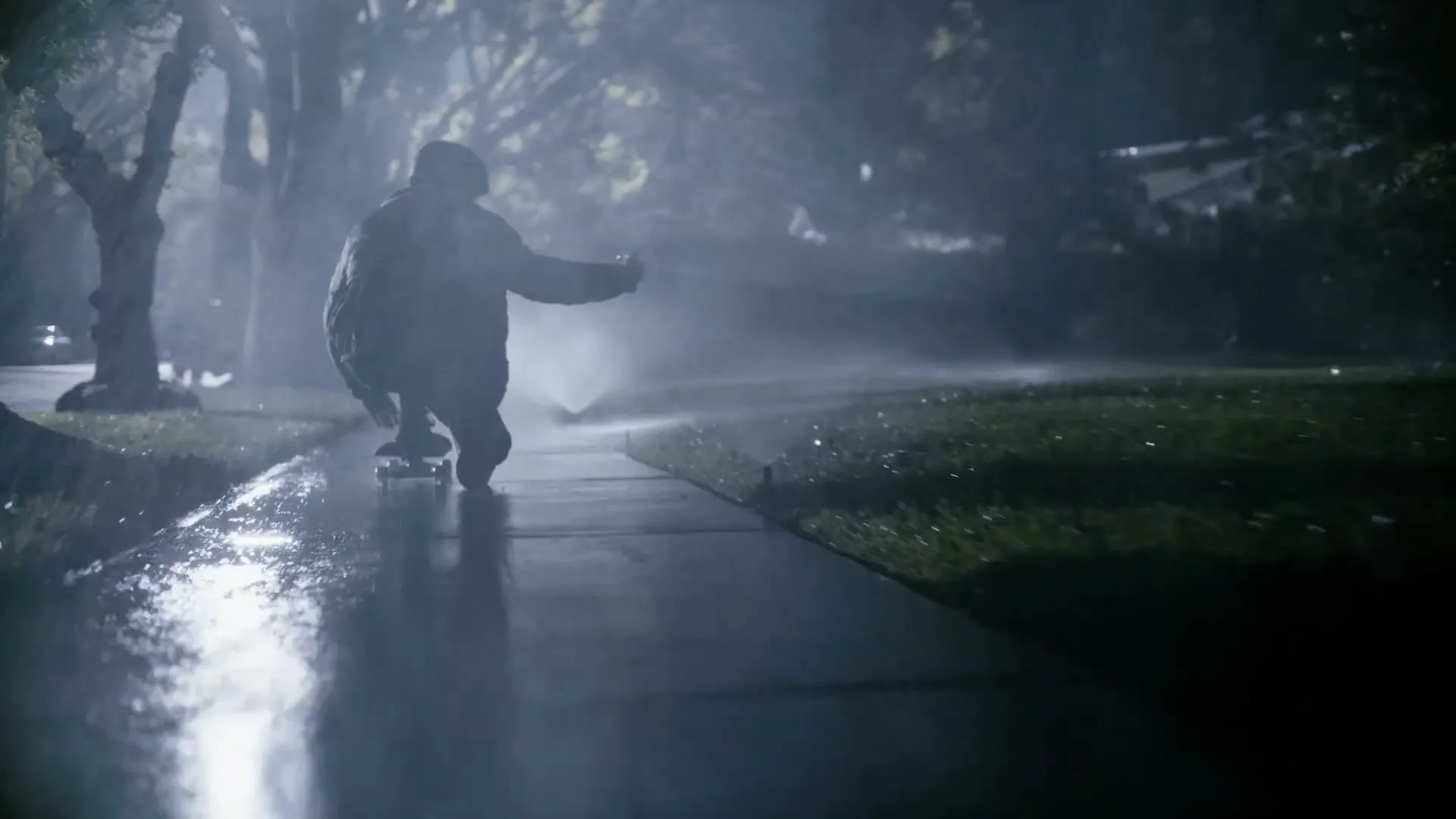 A person in a dark jacket and pants crouches on a skateboard, riding on a wet sidewalk at night, with sprinklers watering the grass.