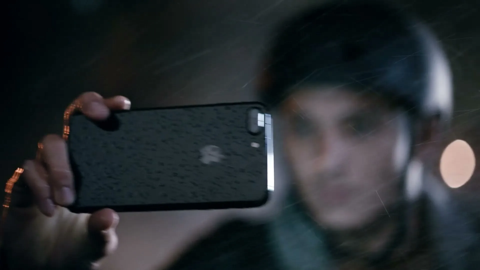 A person holds a wet iPhone horizontally, taking a photo or video in the rain at night, with blurry lights in the background.