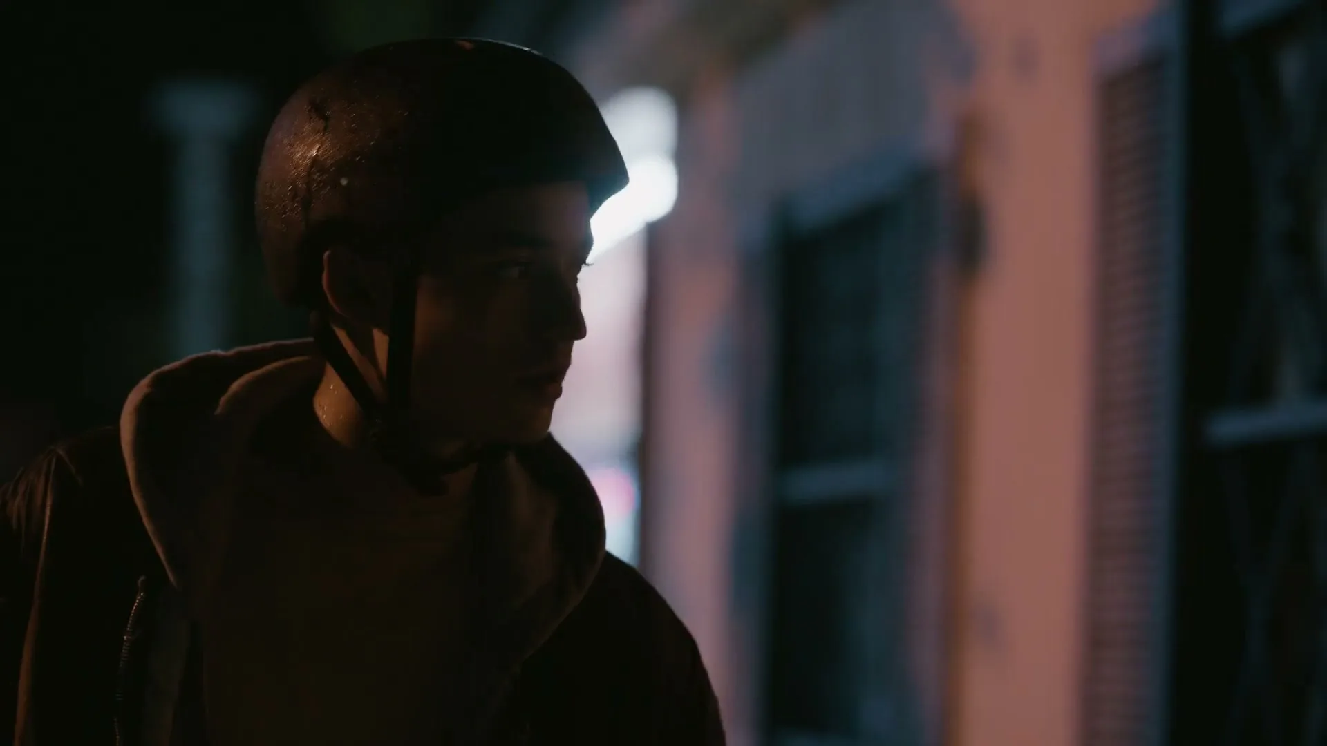 A young man wearing a bike helmet and jacket looks around nervously on a dark street at night.