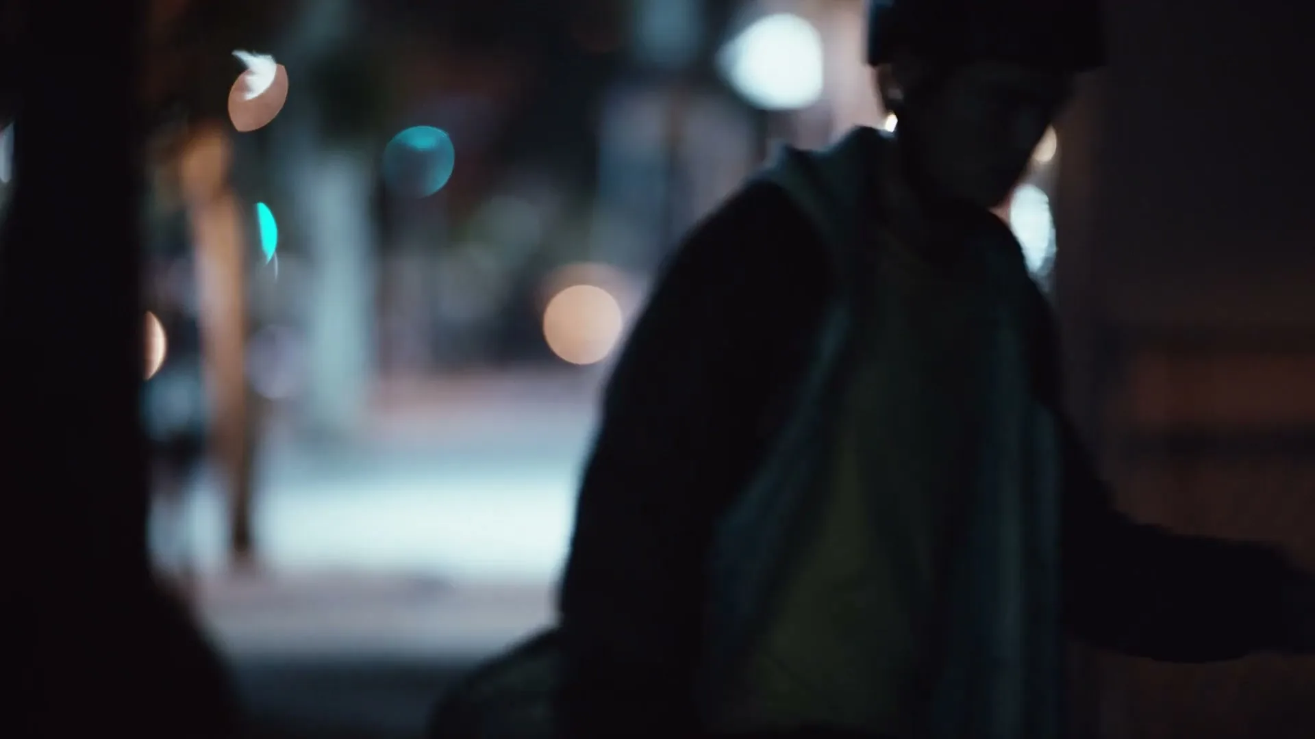 A young man in a yellow shirt and dark jacket walks away from the camera at night, carrying a skateboard under his arm on a city street.