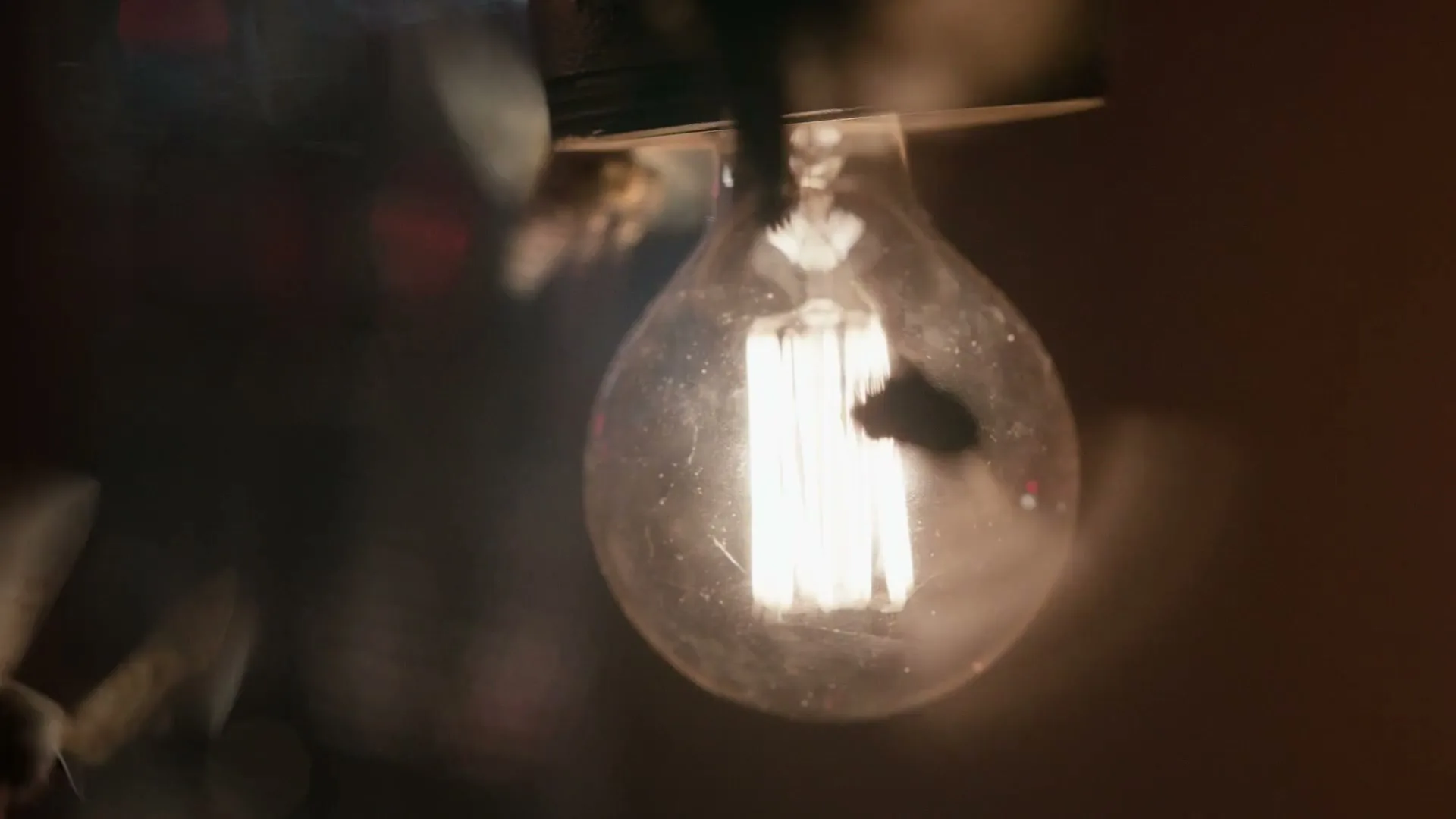 A close-up shot of a single, glowing light bulb with visible filaments, hanging in a dimly lit interior space.