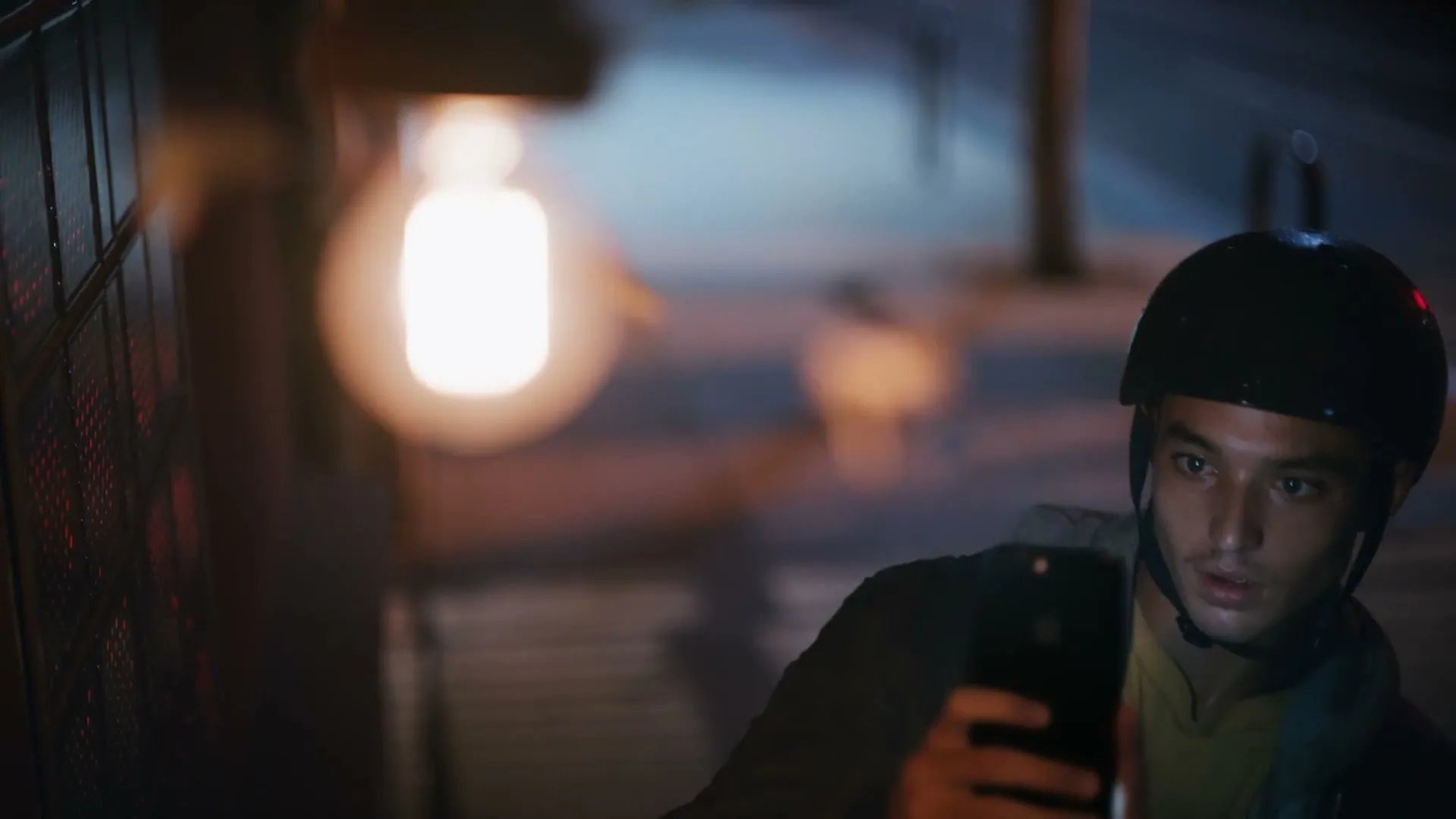 A young man wearing a helmet and a jacket holds a smartphone, looking intently at its screen under a glowing light bulb at night.