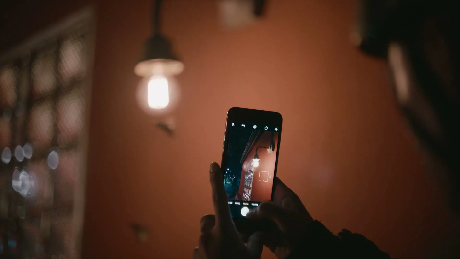 A person holds a smartphone, capturing a photo of a lightbulb hanging from the ceiling in a dimly lit room.