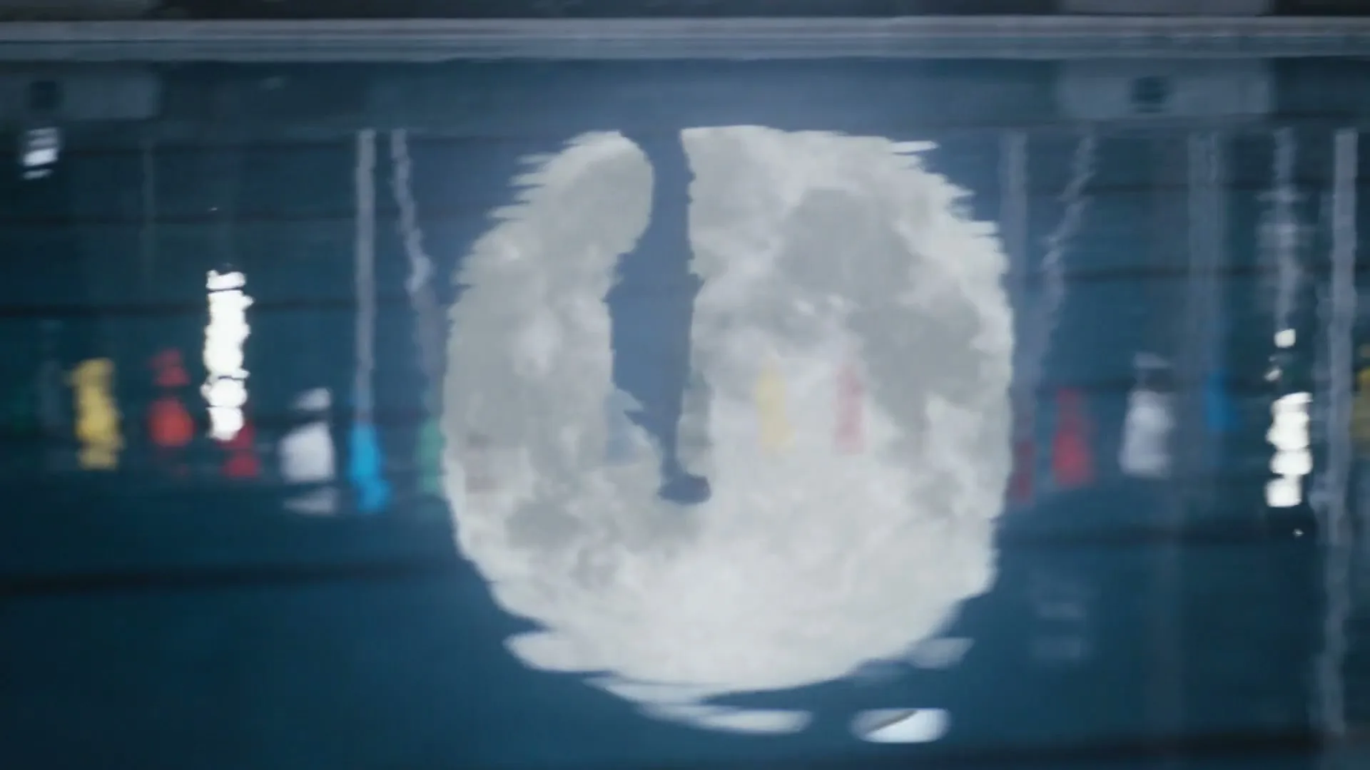 A full moon is reflected on the surface of a swimming pool, with colorful lane markers visible underwater, creating a serene and slightly distorted image.