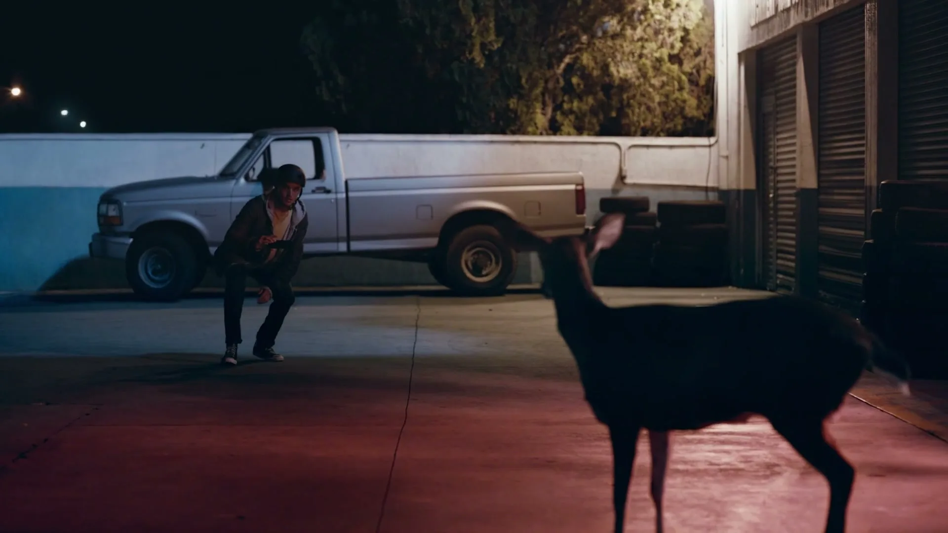 A young man in a hoodie and jeans crouches down to photograph a deer with his phone at night in an industrial area.