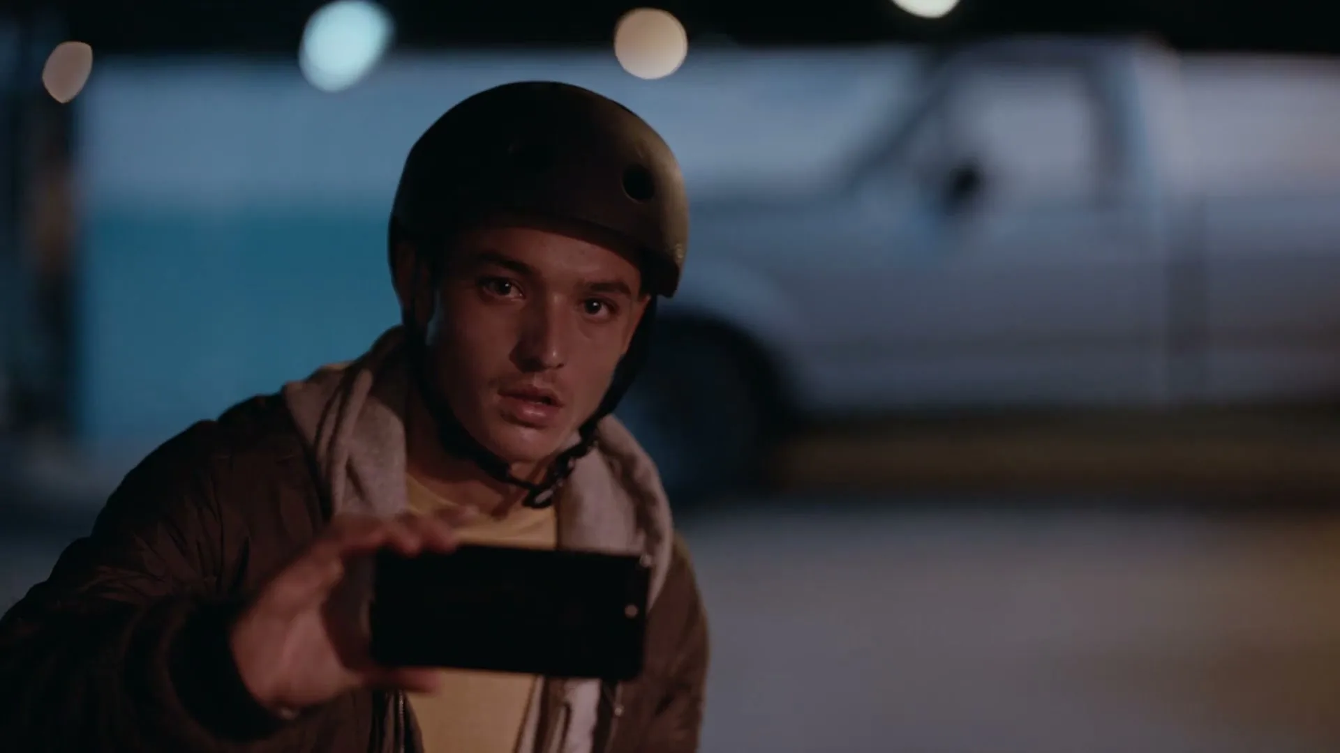 A young male wearing a helmet and jacket holds up a smartphone, looking intently at the screen in a dimly lit outdoor setting at night.