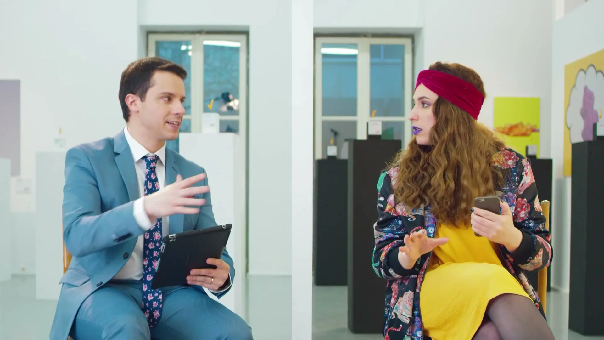 A man in a blue suit and a woman in a yellow dress and colorful jacket sit opposite each other in a modern art gallery, both holding smart devices.