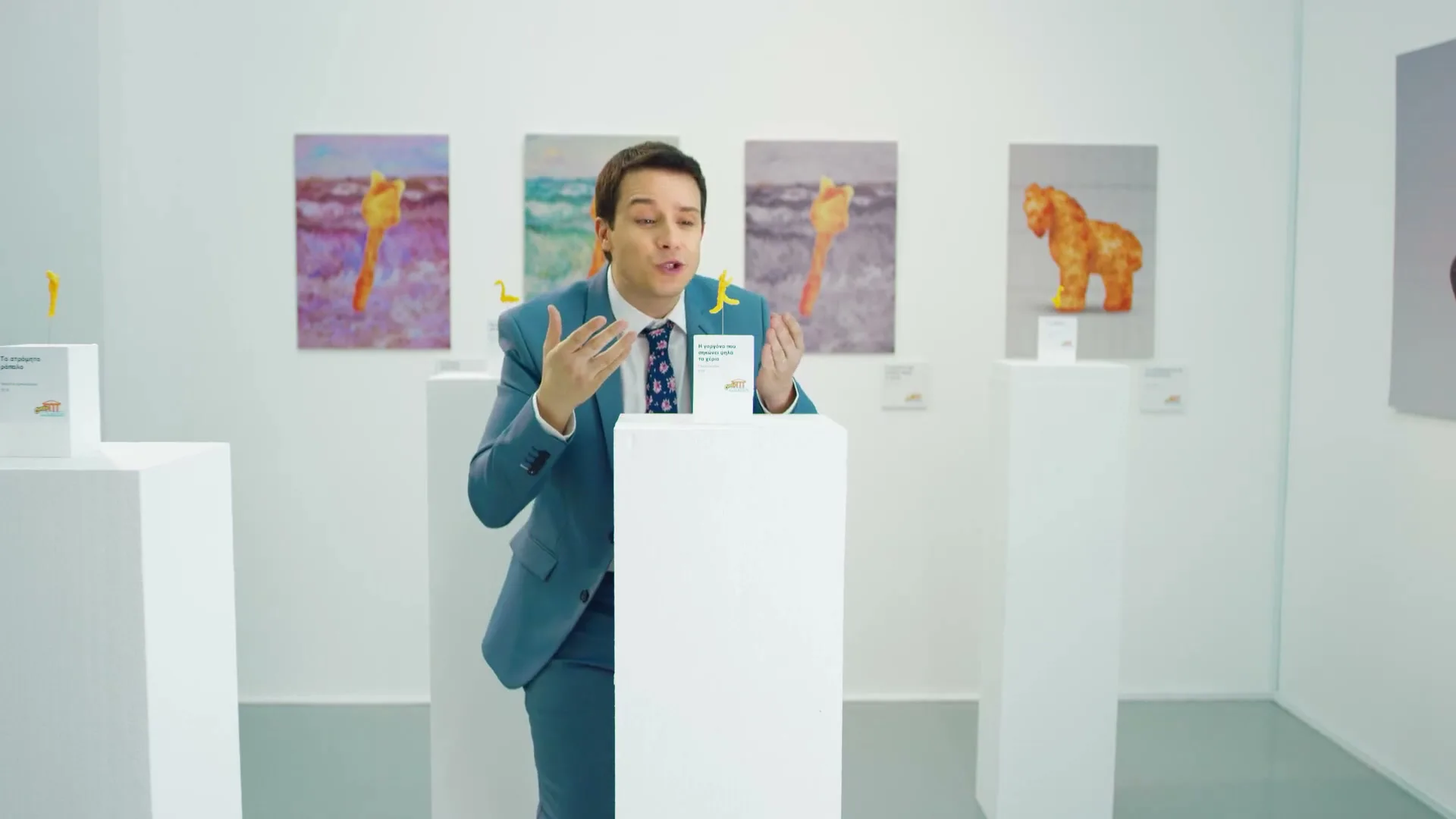 A male art curator in a blue suit stands in a white art gallery, gesturing excitedly about Cheetos-shaped art pieces on pedestals and hanging on the wall.