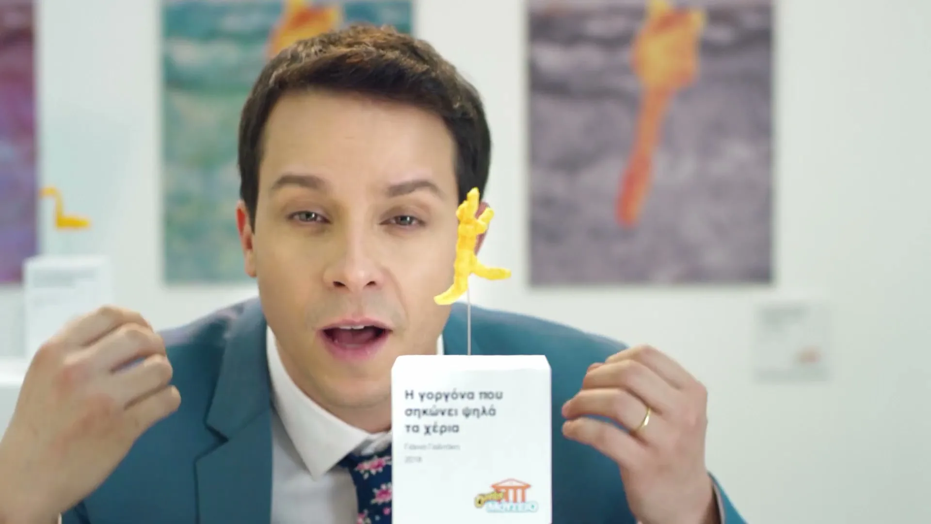 A man in a blue suit and floral tie gestures with his hands while looking at a Cheeto shaped like a mermaid, displayed in a museum.