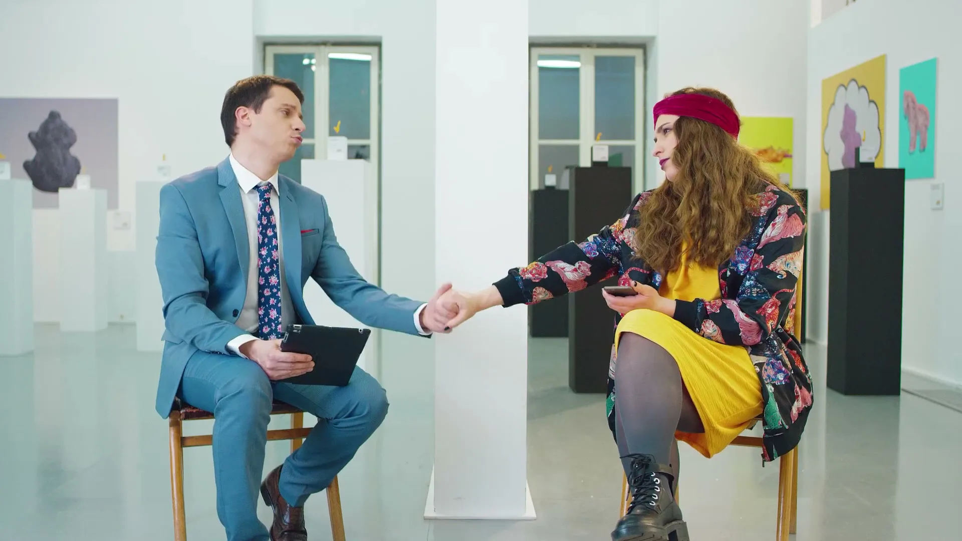 A male host in a blue suit and a female guest in a colorful jacket shake hands in a modern art gallery, both smiling at the camera.