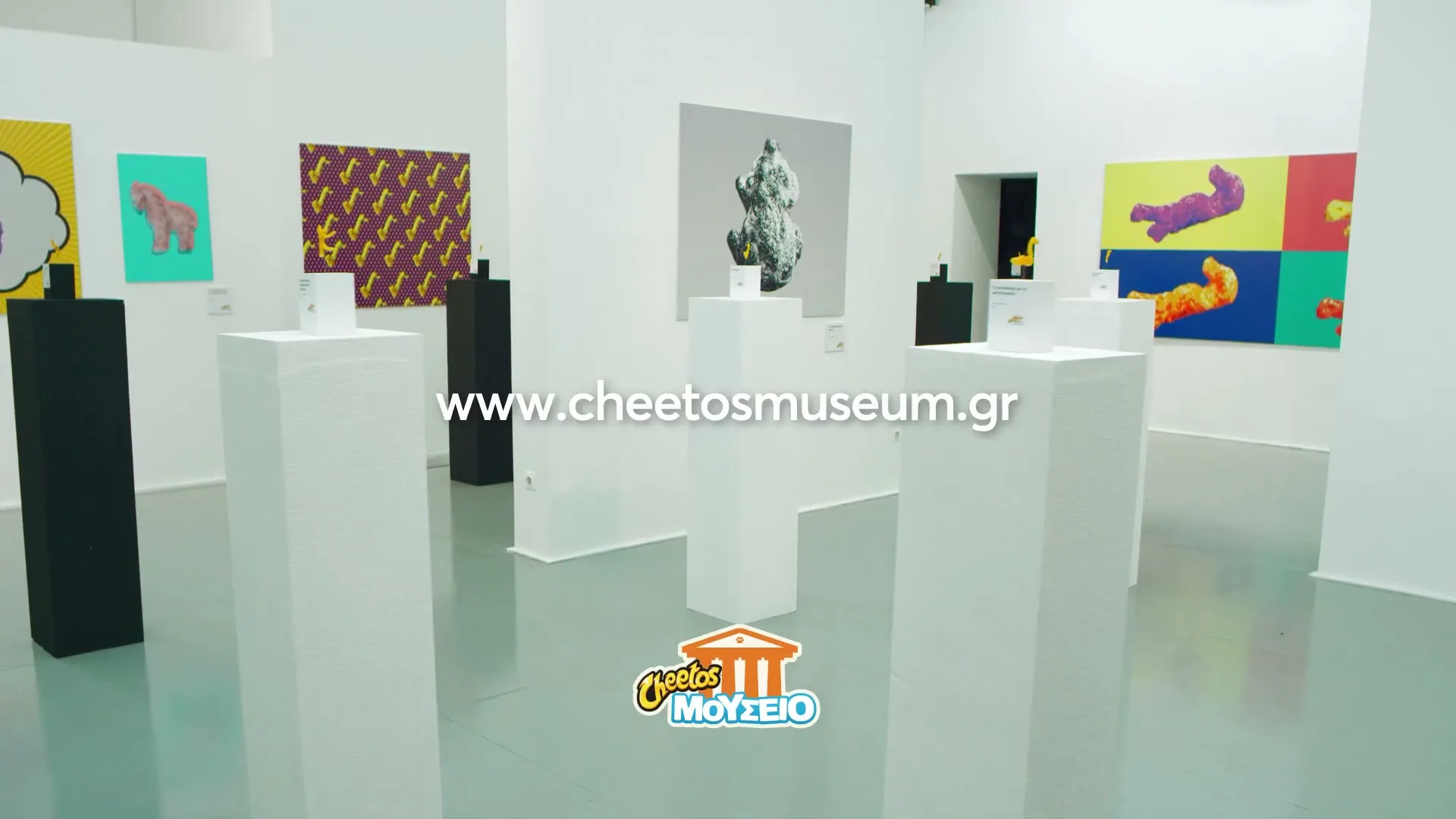 A wide shot of a modern art gallery with white walls and pedestals displaying Cheetos as art, with a website and logo on screen.