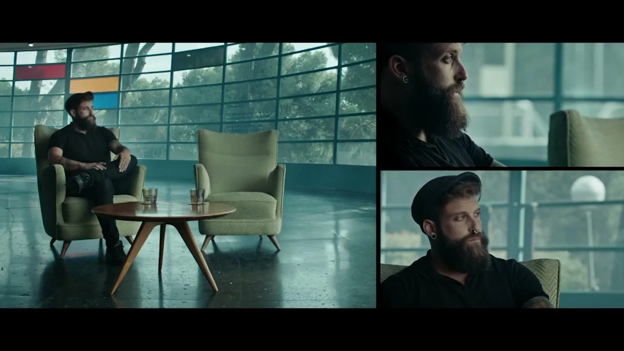 A bearded man in a black shirt and pants sits in a mid-century modern armchair, looking contemplatively out a large window in a modern lounge.