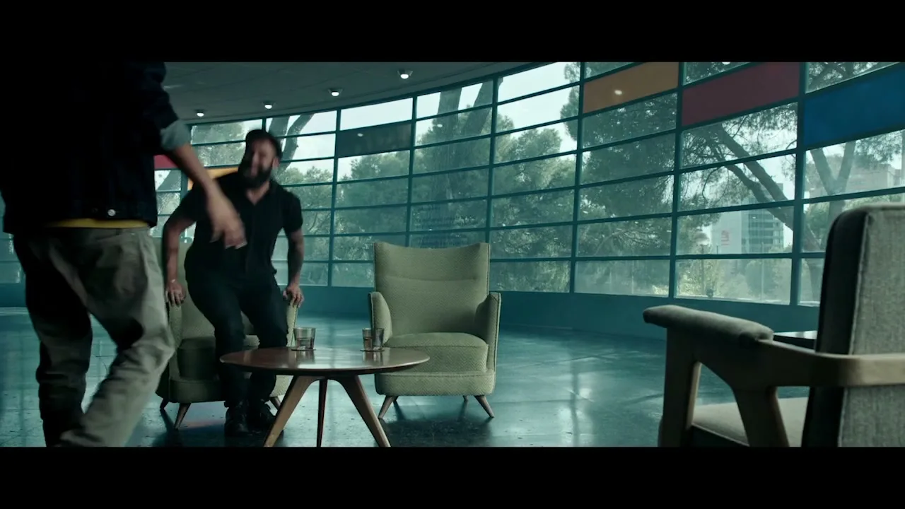 Two men, one with a beard and black shirt, the other in a denim jacket, embrace in a modern lounge with large curved windows overlooking trees.