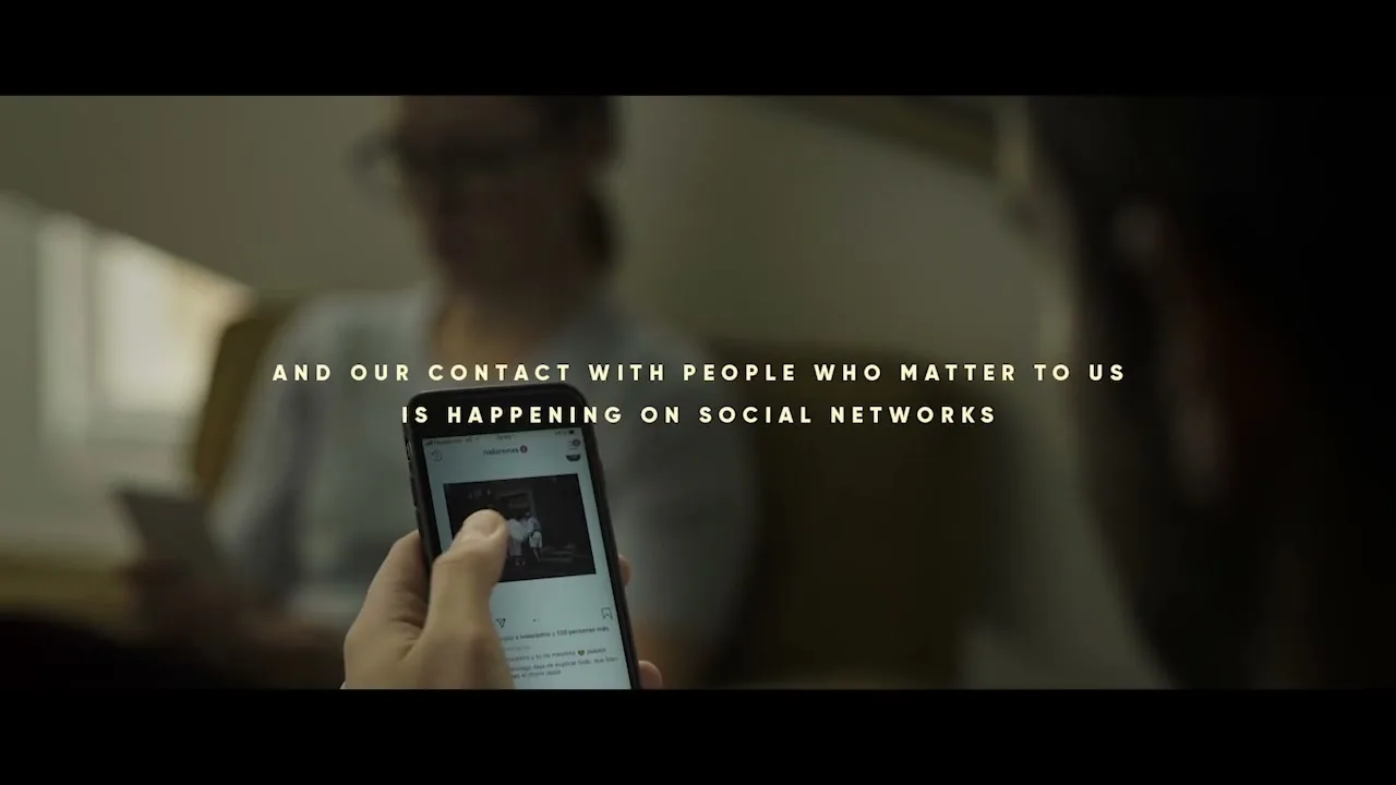 A person's hands hold a smartphone, scrolling through social media, with another person out of focus in the background, in an interior setting.