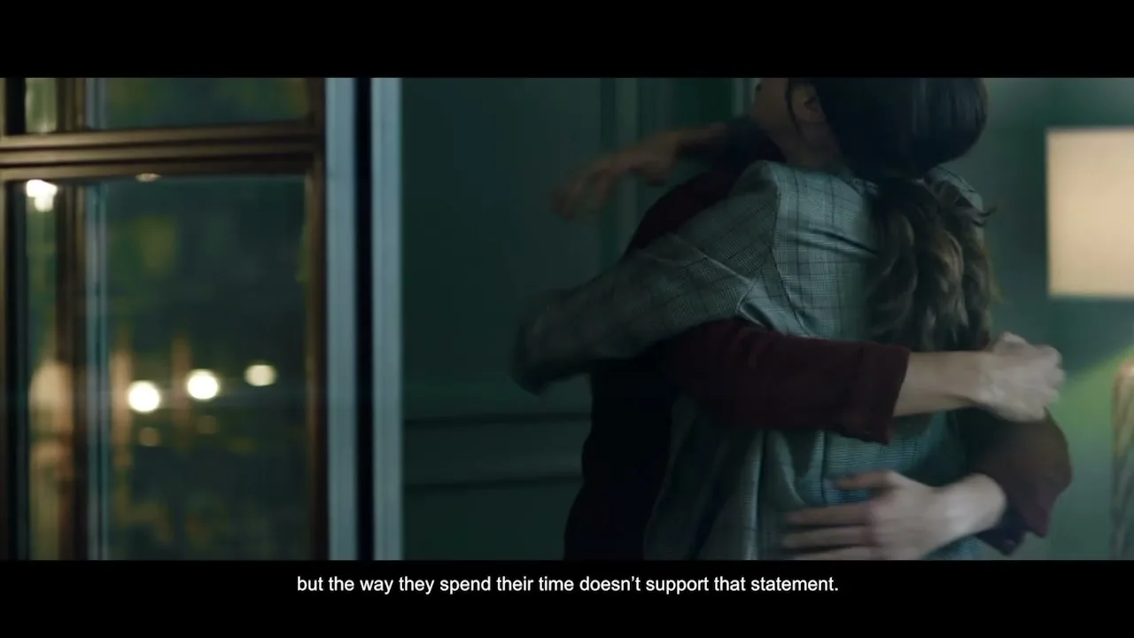 A man and a woman embrace warmly, smiling, in a dimly lit room with a window showing blurred lights outside.