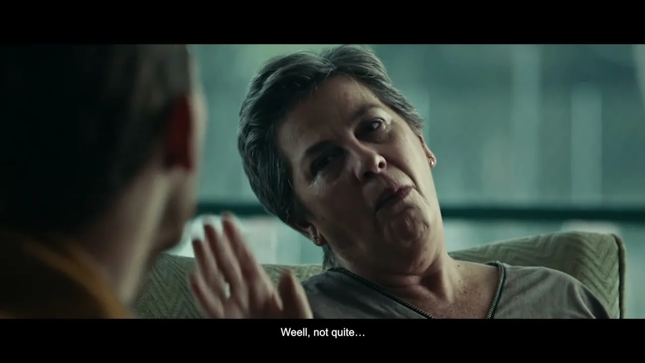 A man with a beard and a woman with short grey hair sit on a couch, the woman touches her face and talks, in an interior setting.