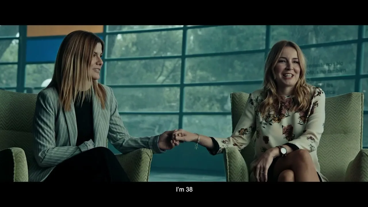 Two women, one in a grey blazer and the other in a floral top, sit in armchairs holding hands and smiling in a modern interior.