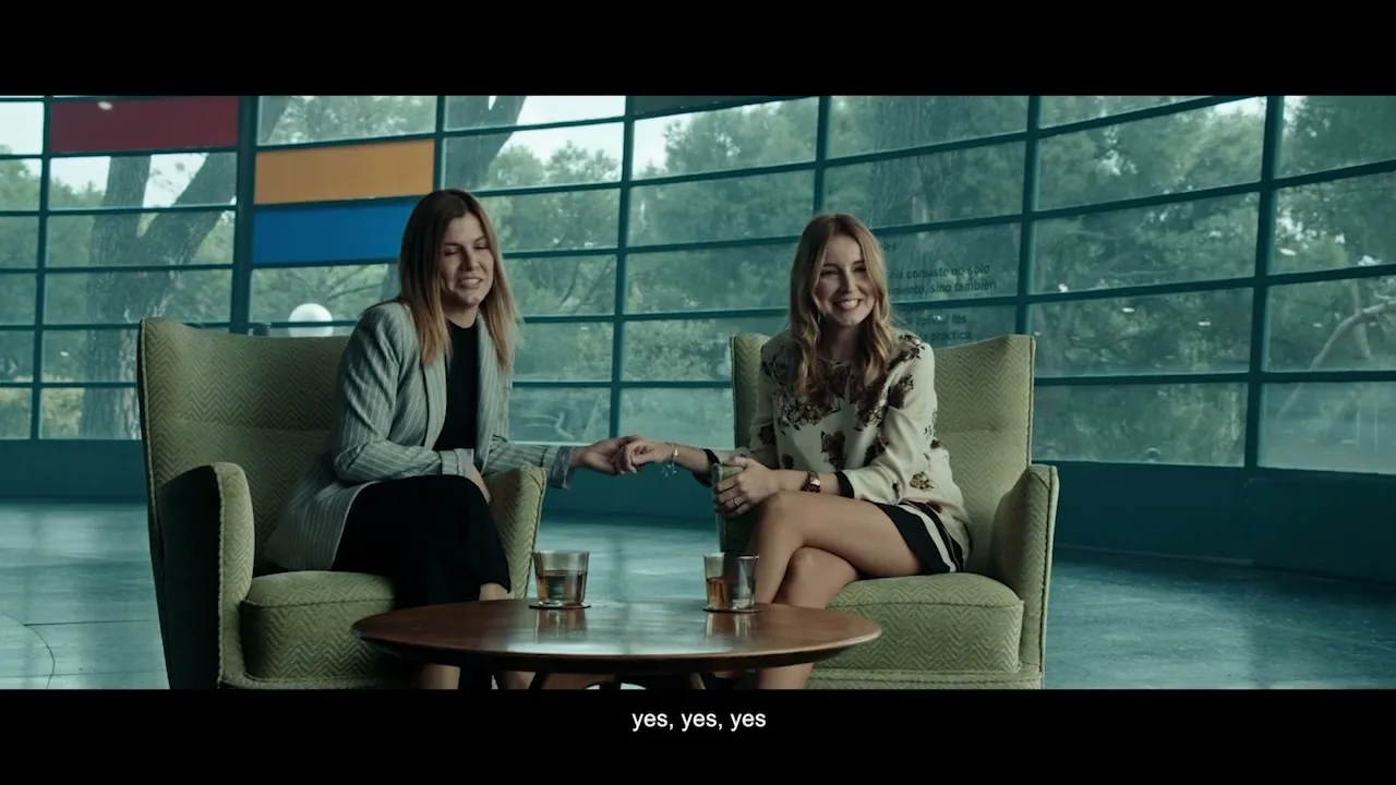 Two women, one in a grey blazer and the other in a floral top, sit in armchairs, holding hands and smiling in a modern building with large windows.