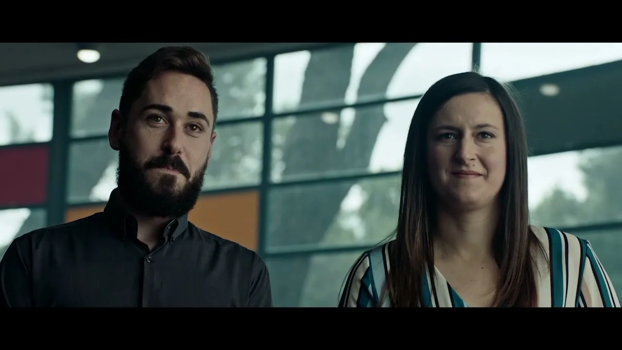 A man with a beard and a woman with long hair stand side-by-side, looking forward and smiling in an office with large windows.