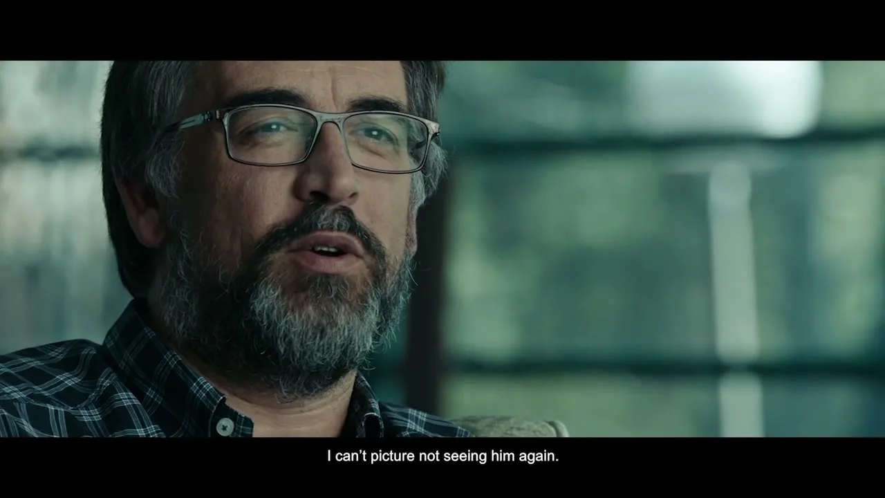 A middle-aged man with glasses and a beard speaks with a somber expression, wearing a plaid shirt in an interior setting.