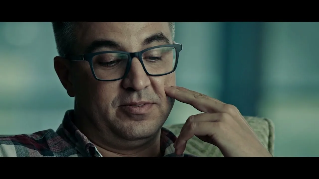 A middle-aged man with glasses and a plaid shirt touches his cheek, looking thoughtful in an interior setting.