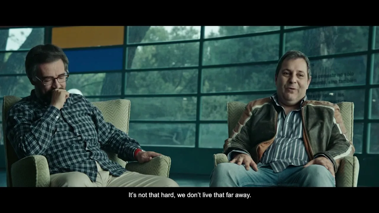 Two men sit in armchairs in a modern room with large windows, one covering his face, the other smiling, as they talk.