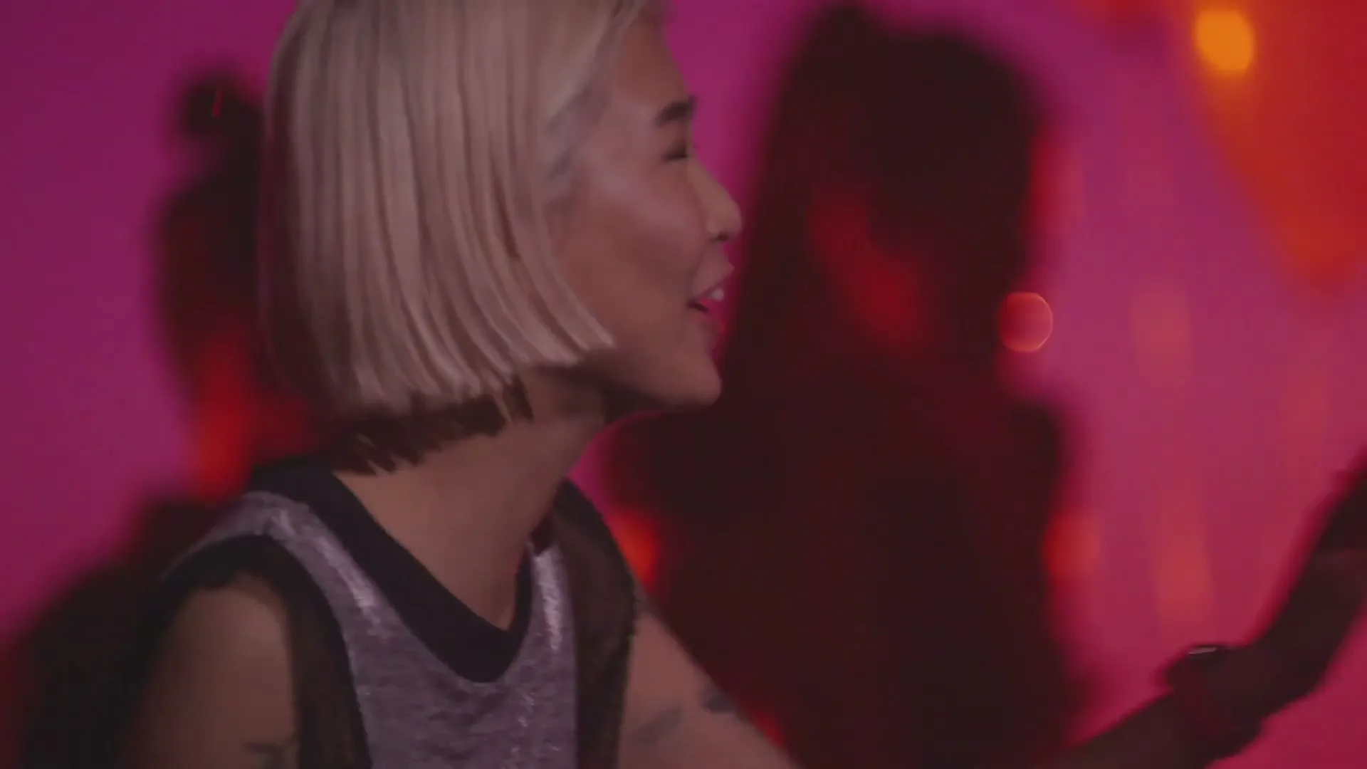 A young woman with blonde hair and a silver top smiles and puts a finger to her lips, shushing someone in a pink-lit room.