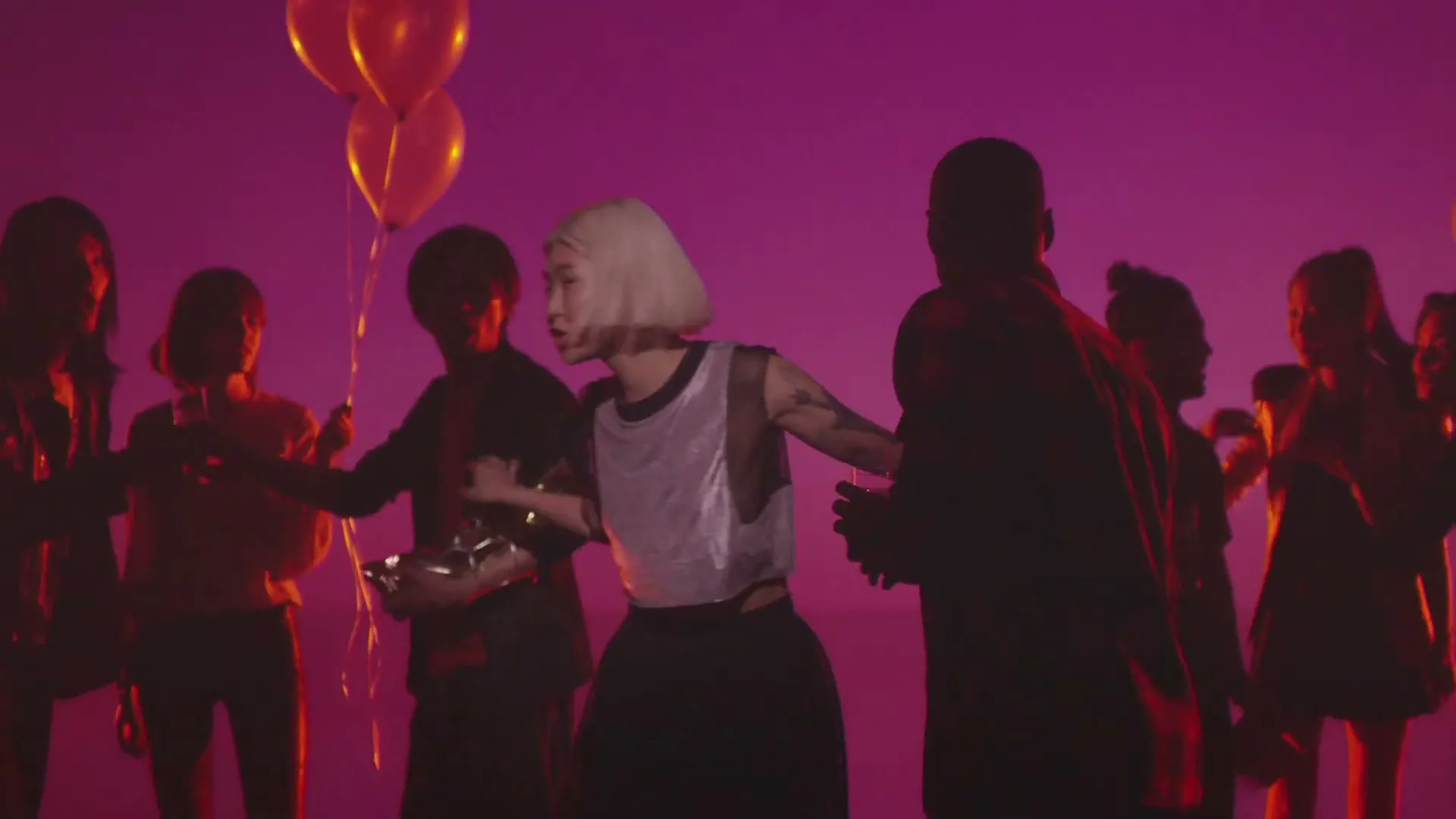 A young woman with blonde hair and a silver top dances with a group of people, holding a gift, in a studio with pink lighting and balloons.