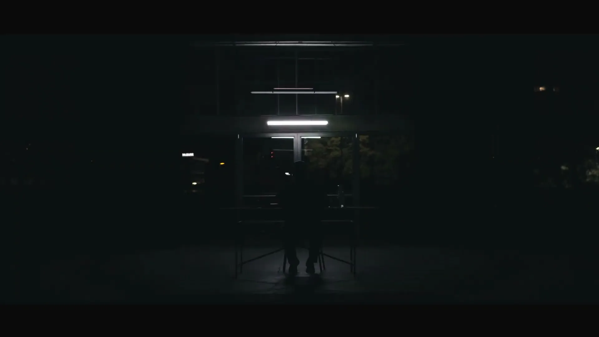 A man sits at a table in a dark, industrial-looking space, illuminated by overhead fluorescent lights, looking directly at the camera.