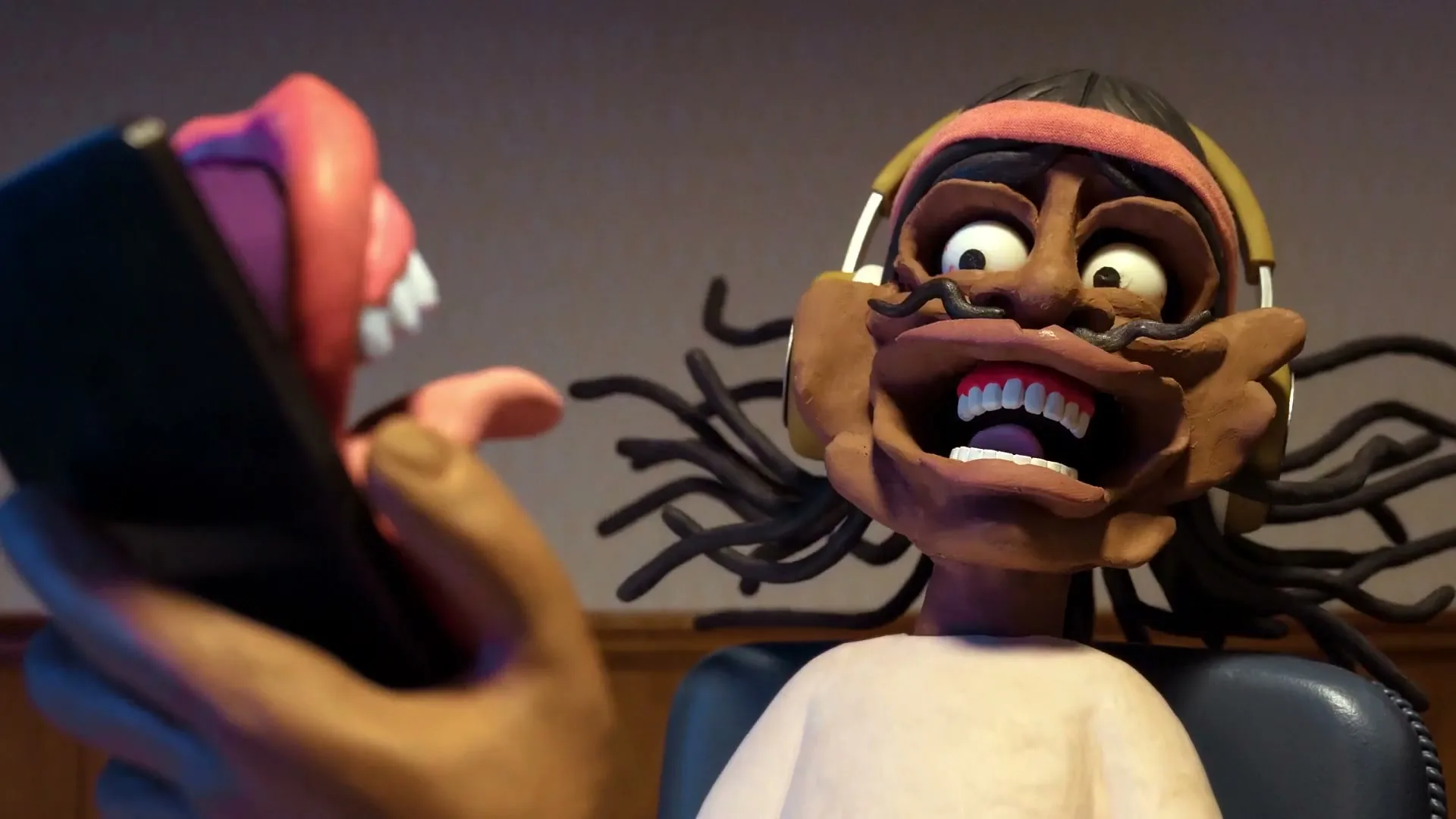 A claymation man with dreadlocks and headphones looks surprised as a pink tongue with teeth emerges from a smartphone he holds.