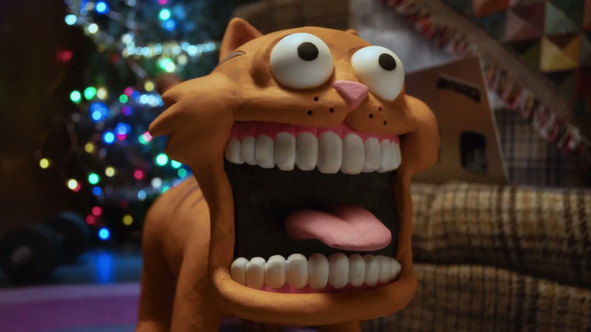 A claymation orange cat with wide eyes and a huge open mouth with teeth and tongue is seen in a room with Christmas lights.