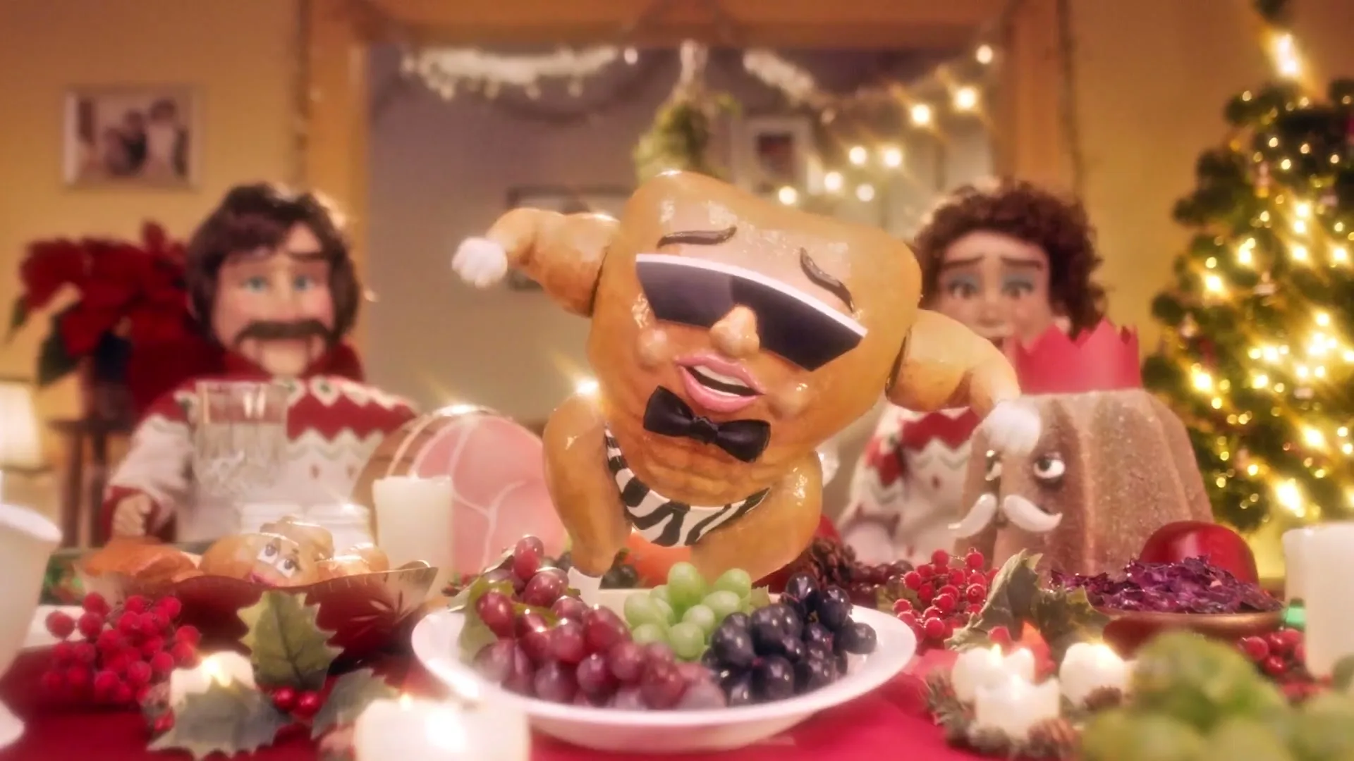 A stop-motion animated turkey wearing sunglasses and a bow tie dances on a festive Christmas dinner table, surrounded by other food items and two puppet-like figures.