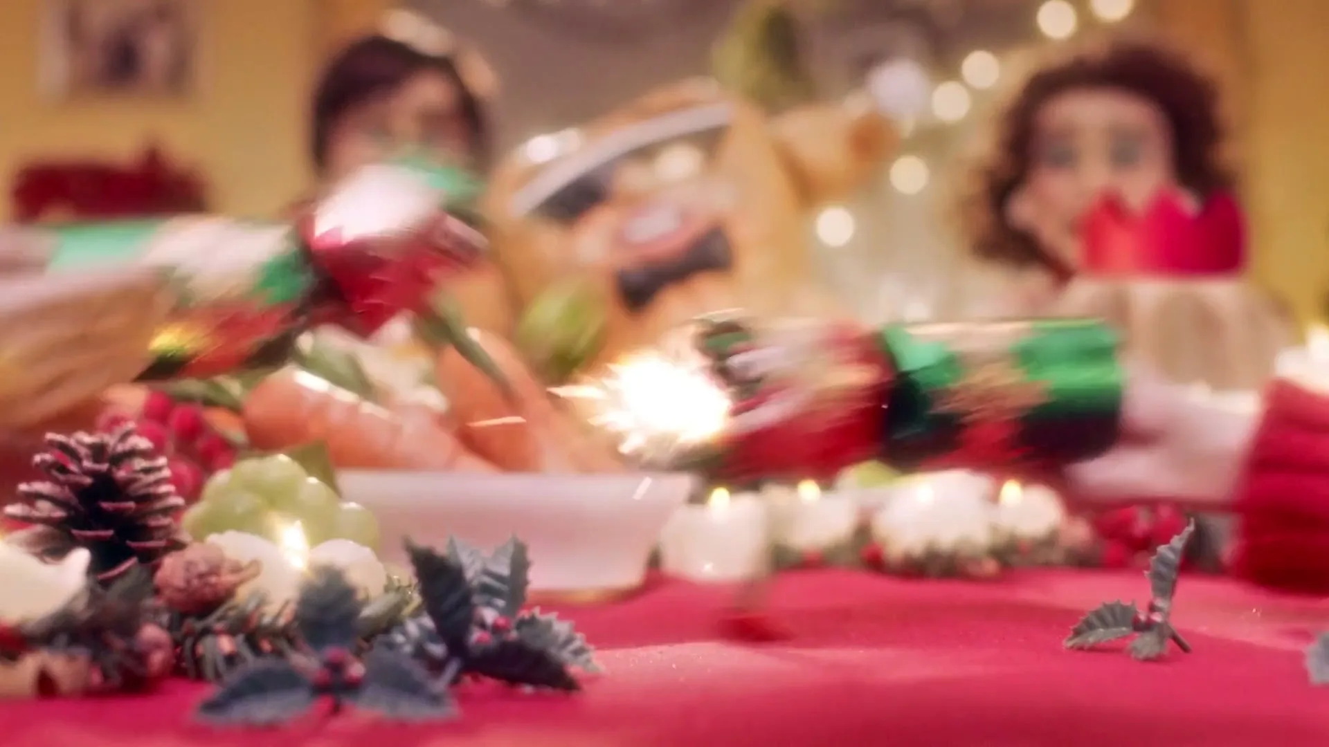 Two pairs of hands pull a festive Christmas cracker with a face and legs, causing it to pop and spark, on a red table with blurred figures in the background.
