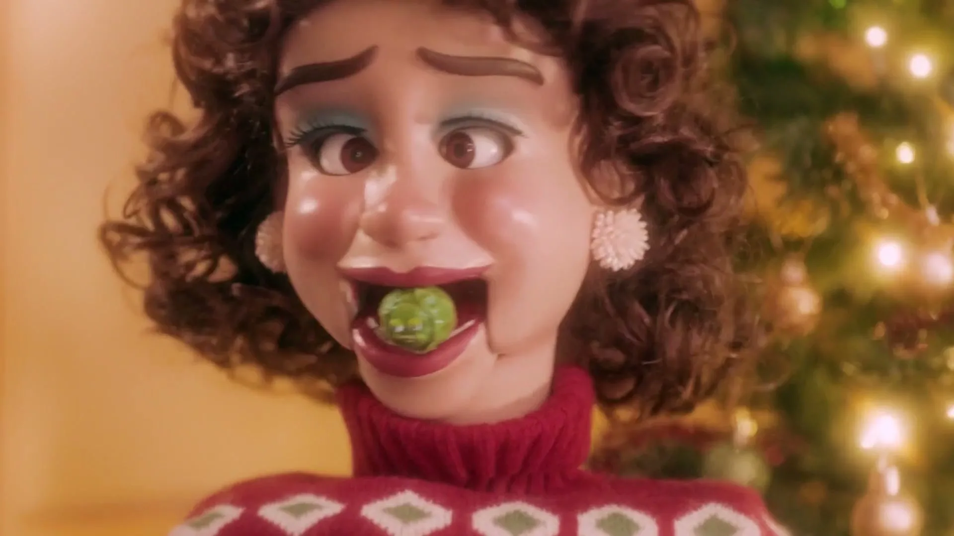 A female puppet with curly brown hair and a red sweater has a small green creature in her mouth, in front of a Christmas tree.