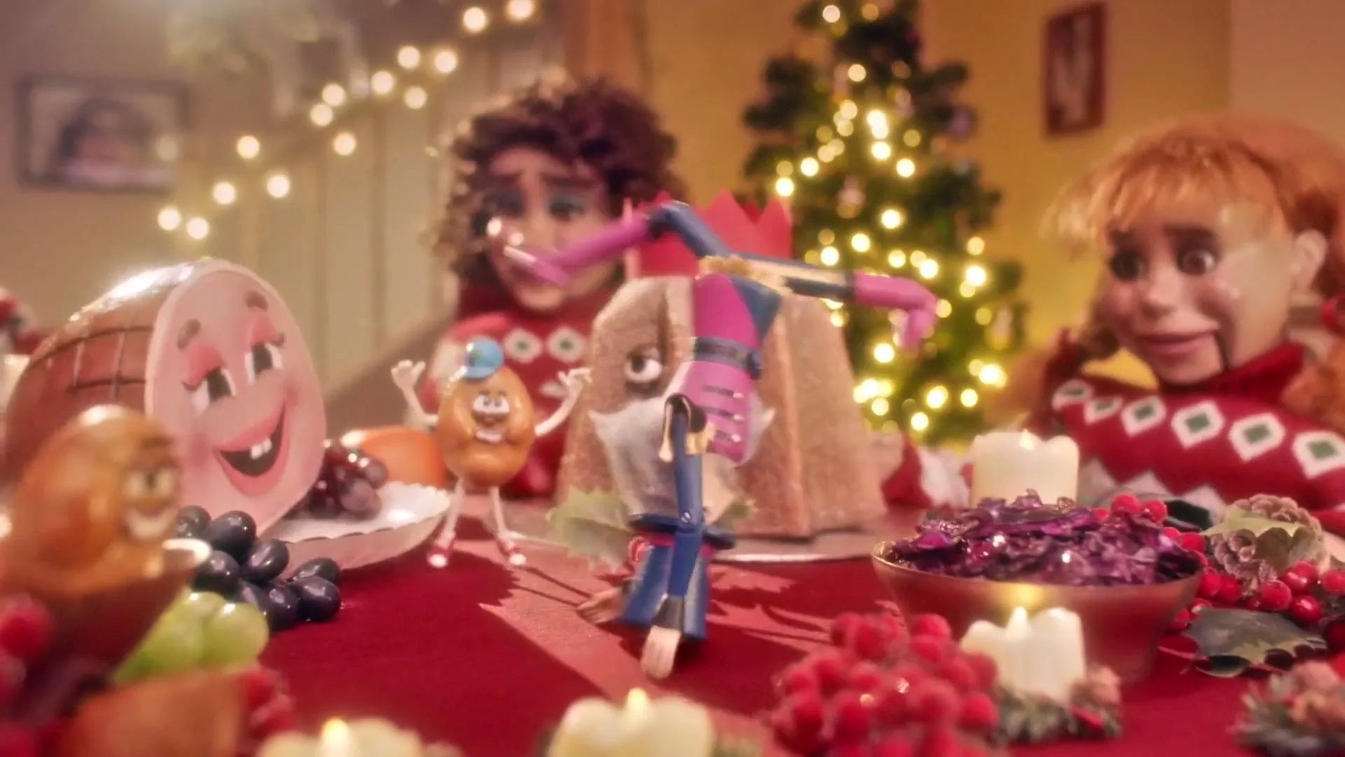A stop-motion animated Christmas dinner scene features a nutcracker dancing on a table surrounded by food characters and two puppet-like figures.