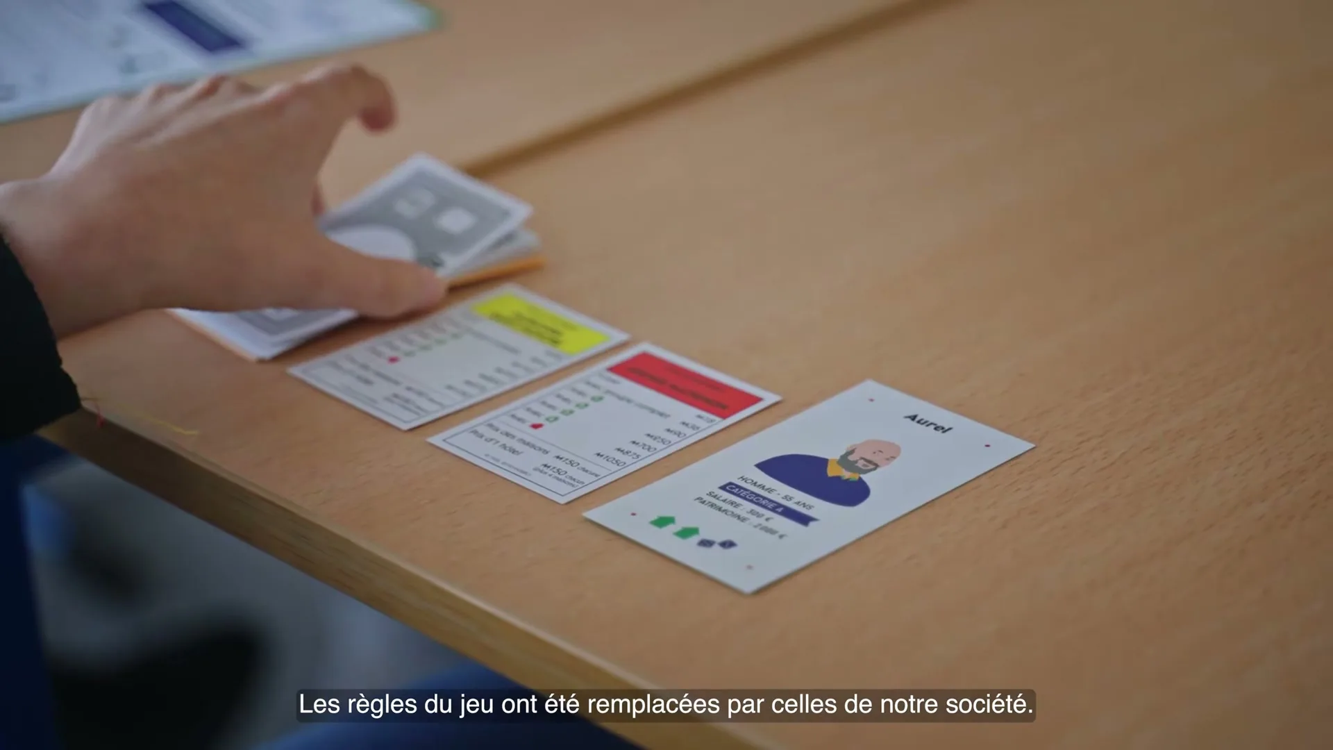 A person's hand places a game card on a wooden table next to other game cards, including one with a cartoon man named Aurel.