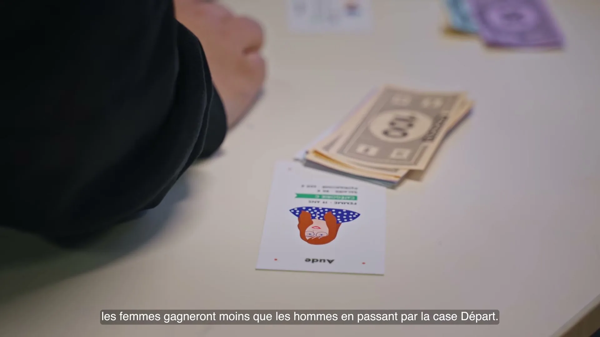 A person's arm is visible as they play a board game with cards and money on a white table, with a character card featuring a woman named Aude.