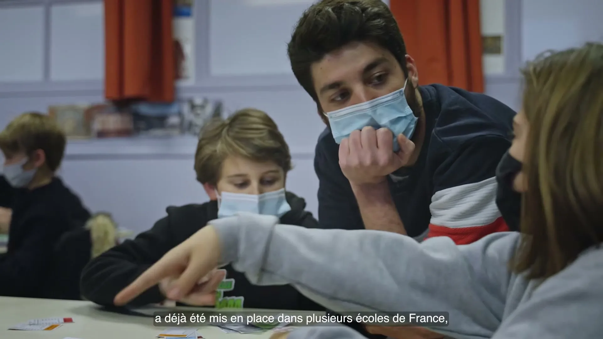 A male teacher and two students, all wearing face masks, are seated at a table in a classroom, looking at something on the table.