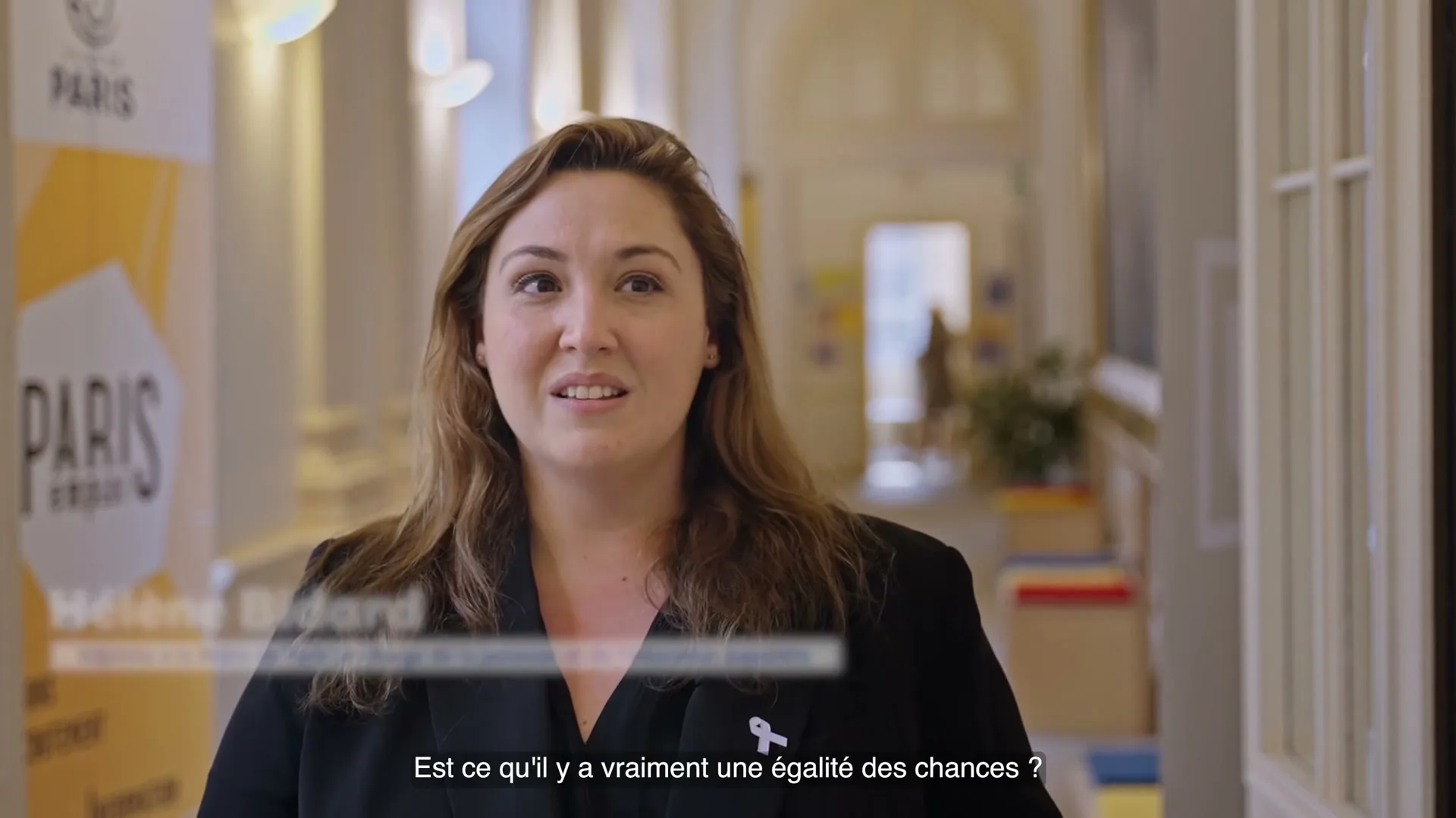 A woman with long brown hair and a black blazer speaks directly to the camera in a well-lit hallway, with a "Paris" banner visible in the background.