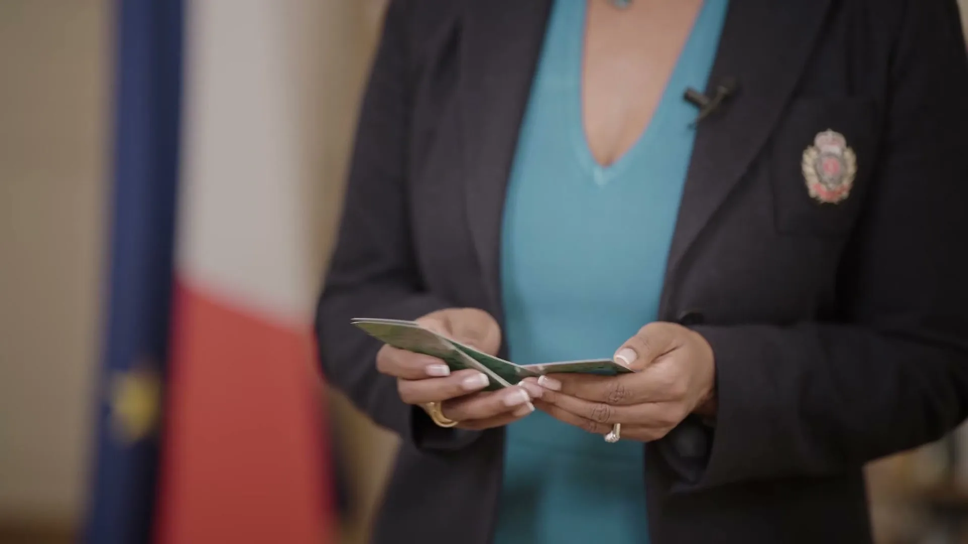 A woman in a black blazer and blue top holds and shuffles green cards, possibly game cards, indoors with a French flag in the background.