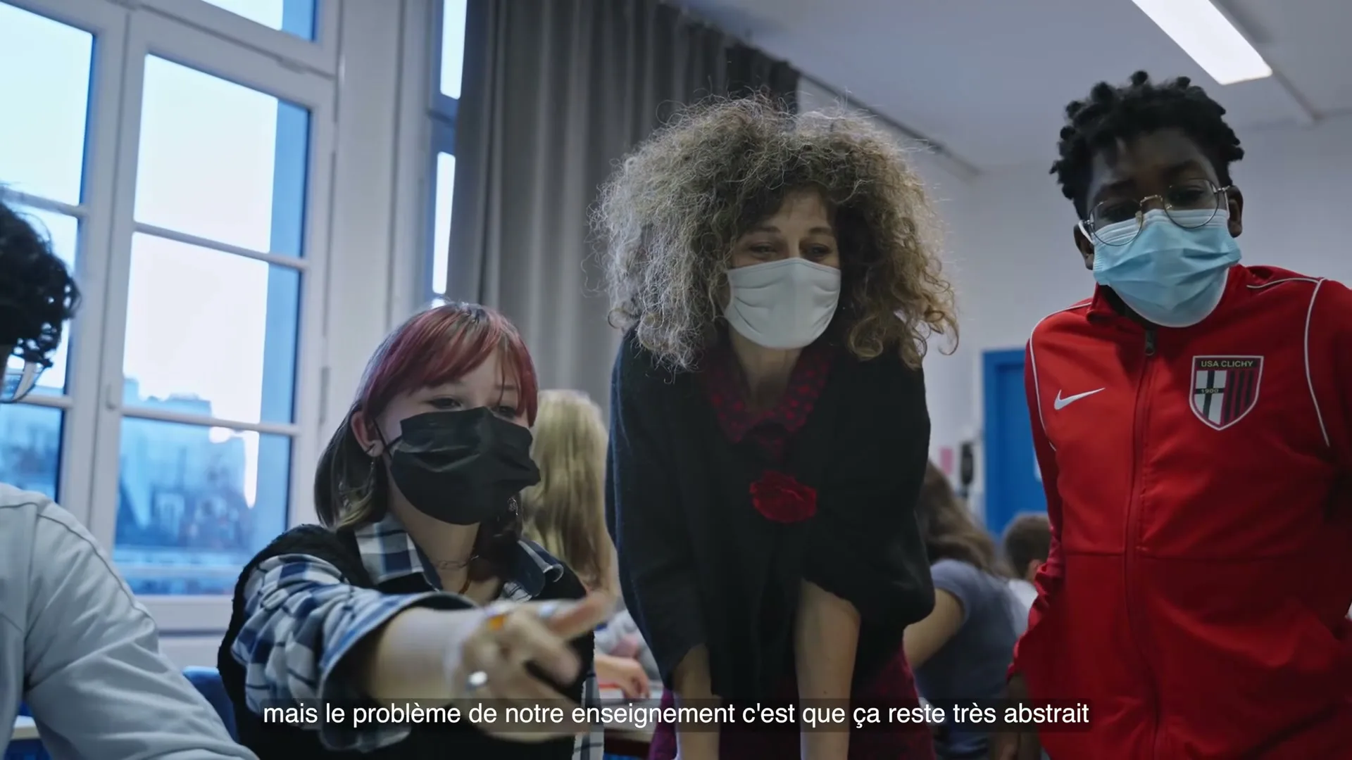 A diverse group of masked students and a teacher in a classroom, with the teacher leaning over a desk, engaging with the students.