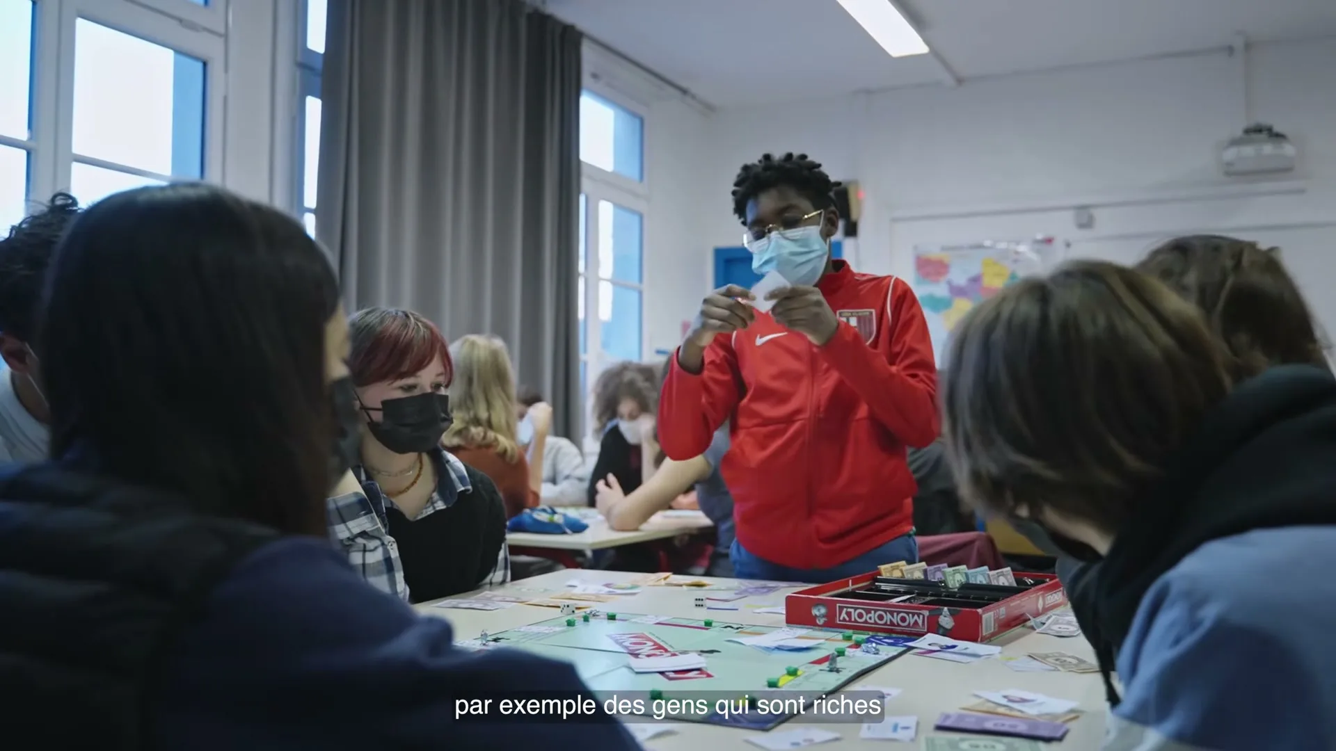 A group of diverse teens in face masks play Monopoly in a classroom, with one teen in a red jacket holding up game money.