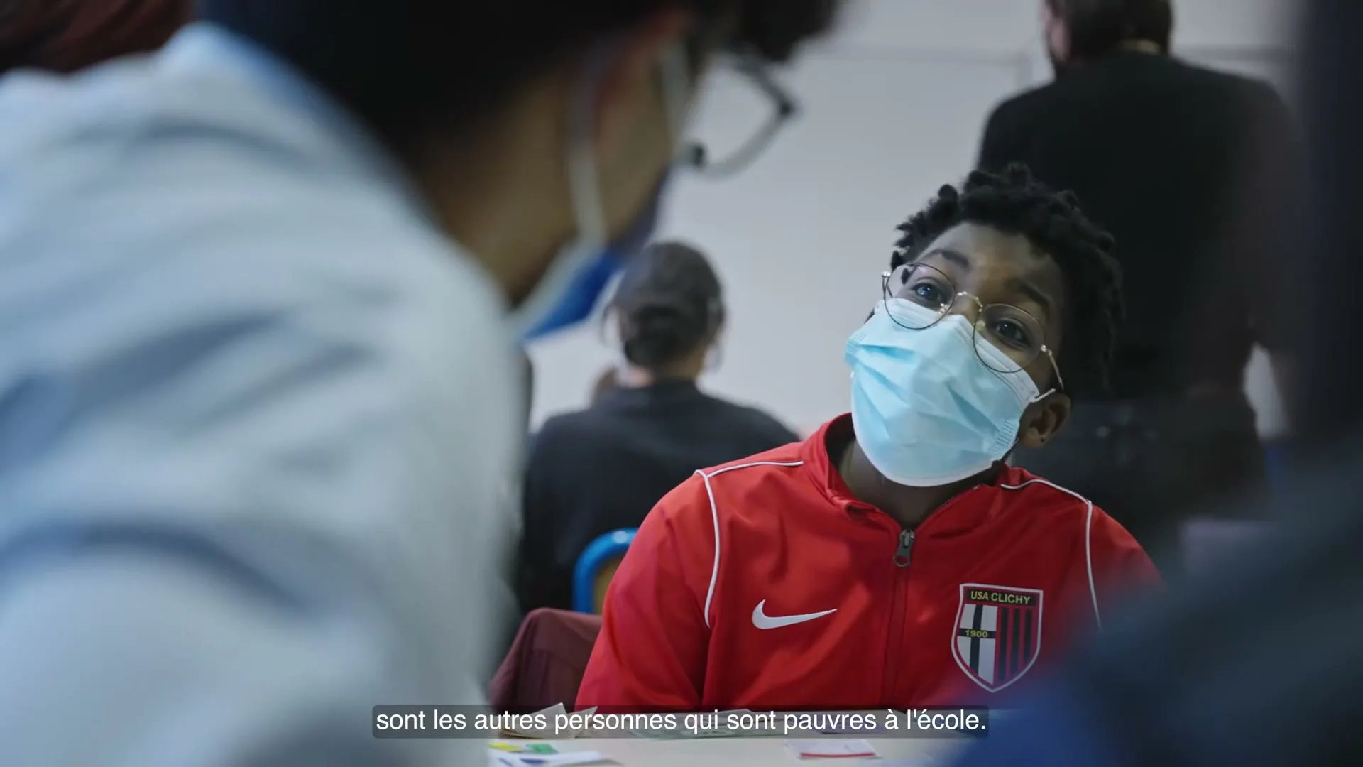 A young boy in a red jacket and face mask looks up at someone off-camera while sitting at a table in a classroom.