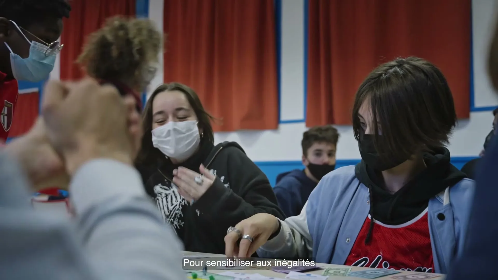 Two masked teens play a board game at a table in a classroom, with other students visible in the background.