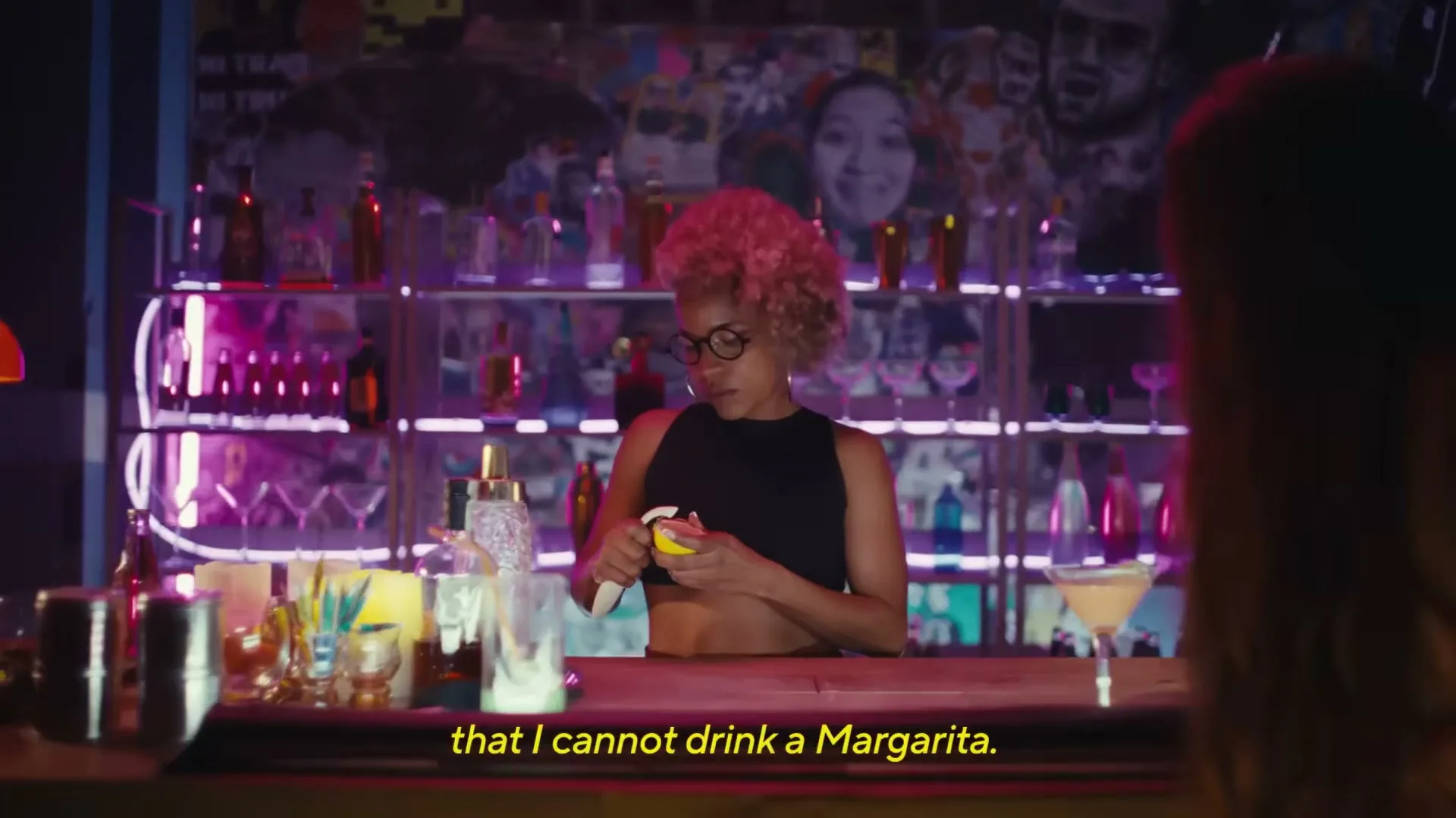 A female bartender with pink curly hair and glasses squeezes a lemon behind a neon-lit bar, with other patrons visible in the foreground.