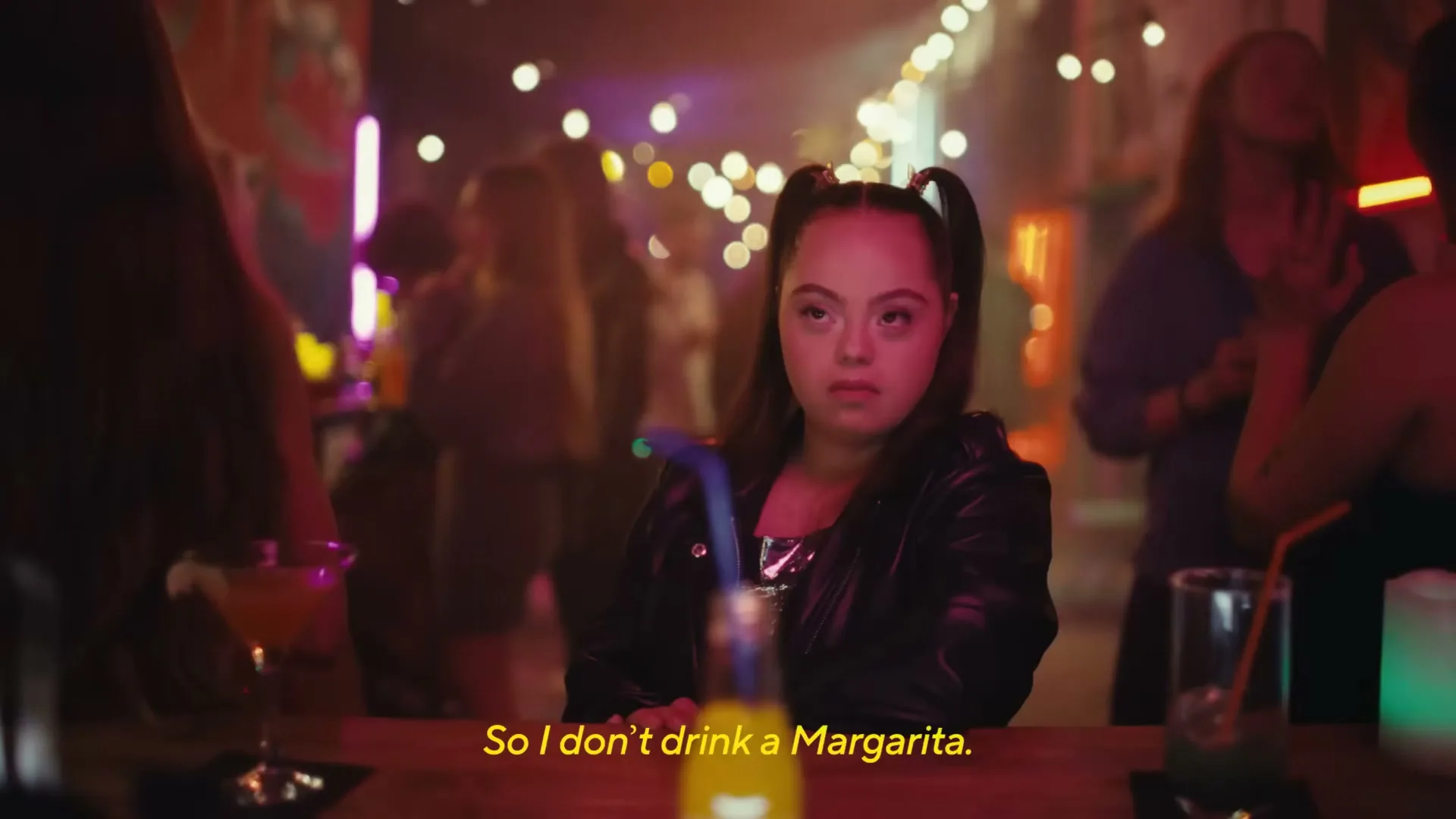 A young woman with Down syndrome sits at a bar, looking directly at the camera with a serious expression, surrounded by blurred people in a dimly lit club.
