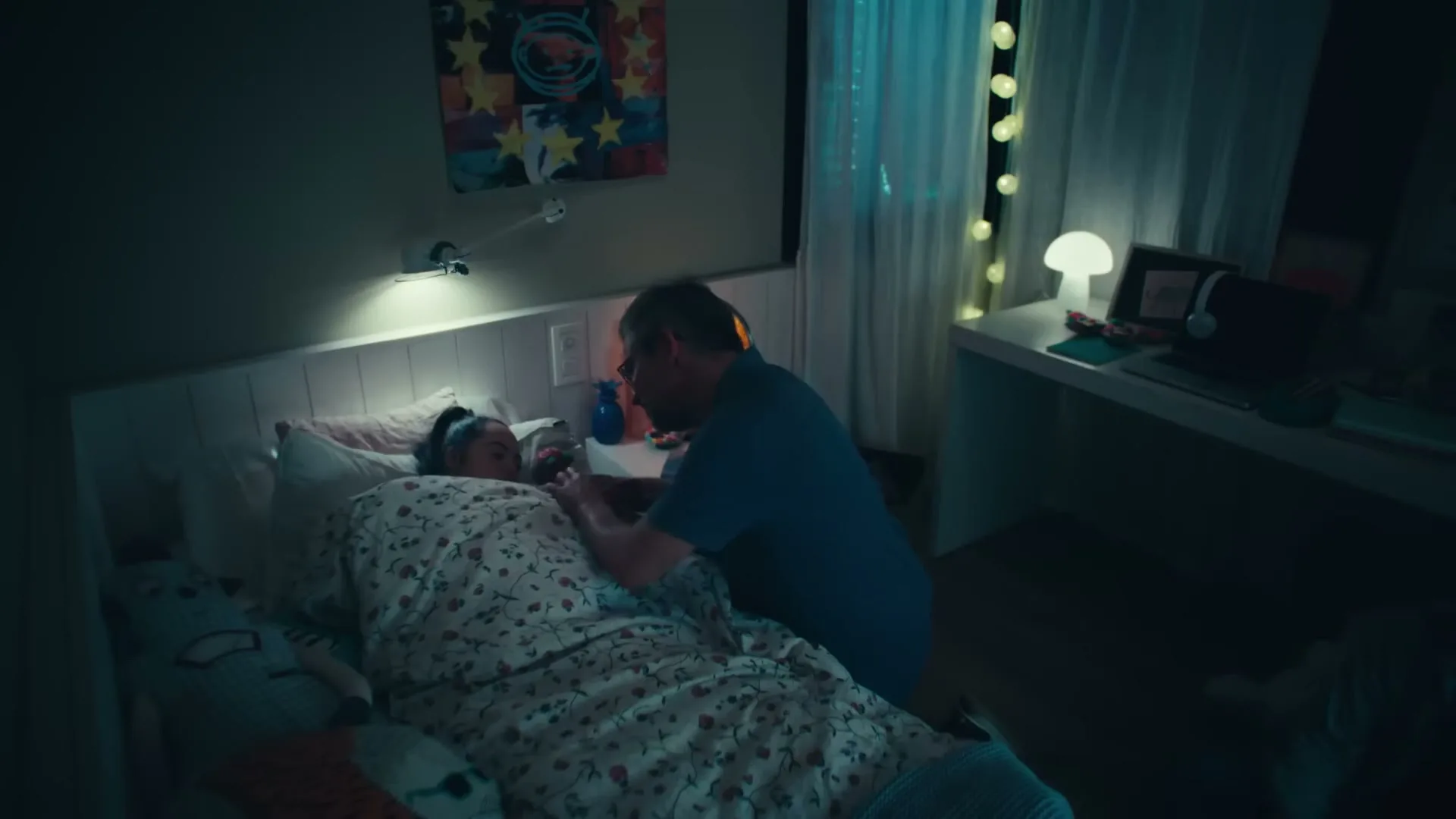 A father in a blue shirt tucks his daughter with Down syndrome into bed in her bedroom at night, gently patting her arm.