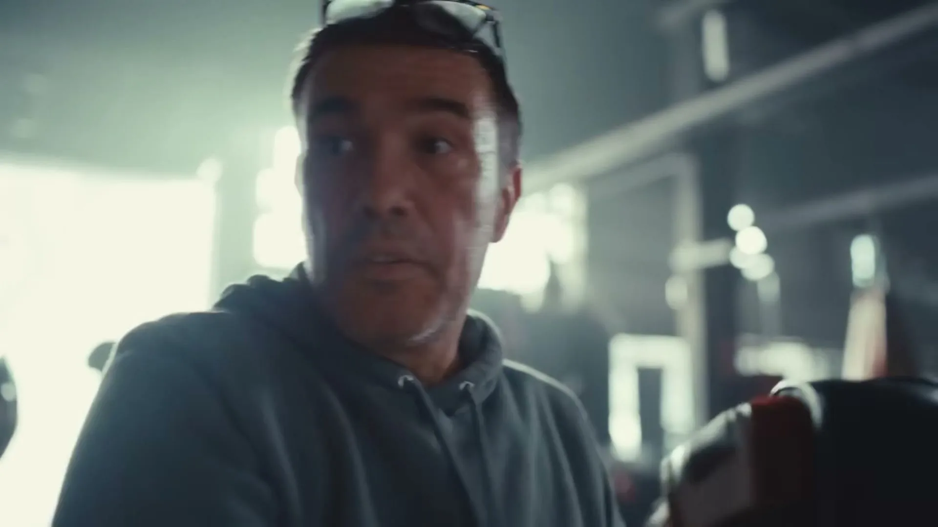 A man with dark hair and a grey hoodie looks around with a serious expression in a dimly lit gym.